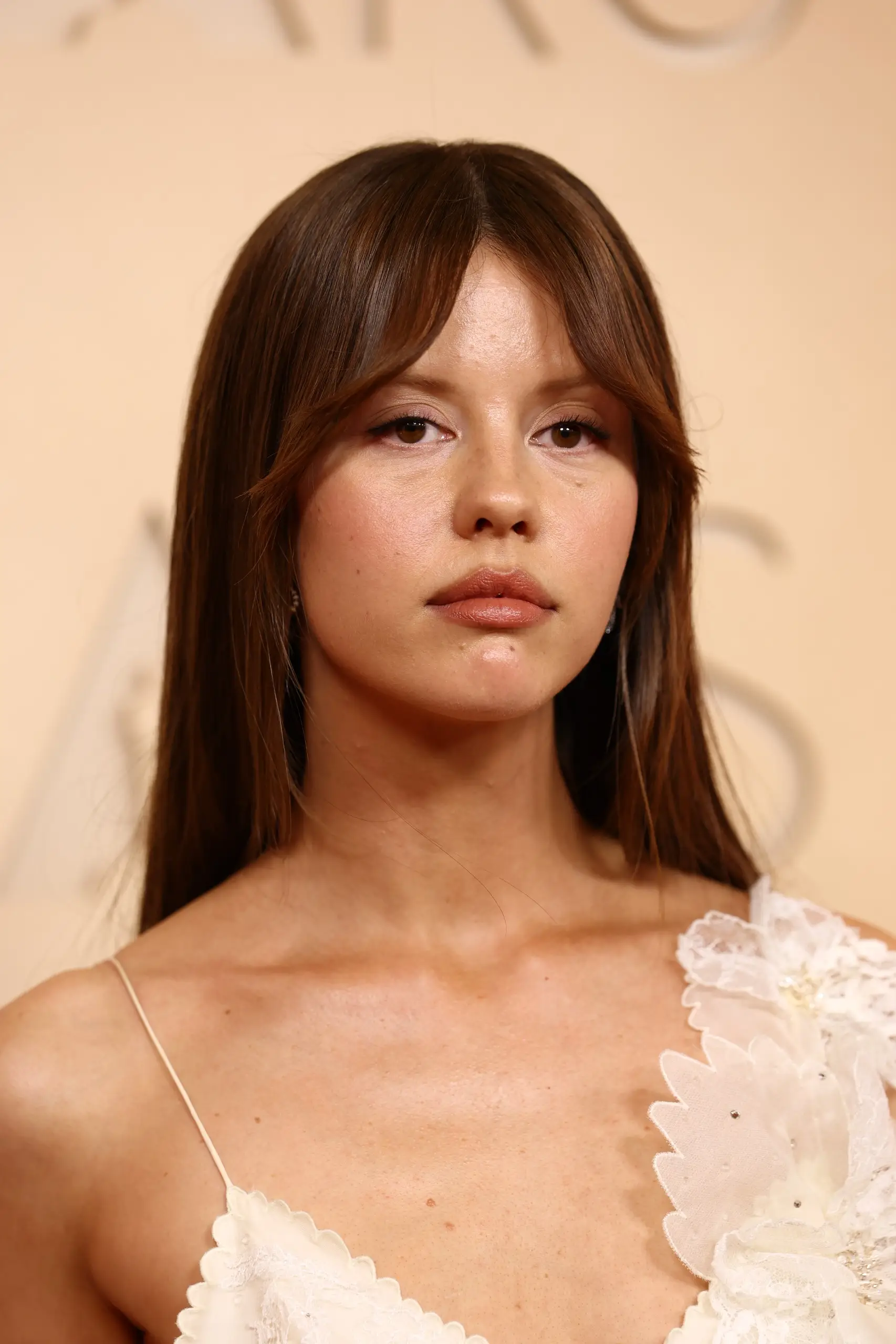 Mia Goth, uzun kahverengi saçları ve alnına düşen perçemleriyle, hafif makyajlı yüzü ve dikkat çekici kahverengi gözleriyle kameraya doğrudan bakarken, zarif fildişi rengi, omuzunda işlemeli dantel detaylara sahip elbisesiyle 2026 Oscar töreniyle ilgili bir etkinlikte şık bir şekilde görülüyor.