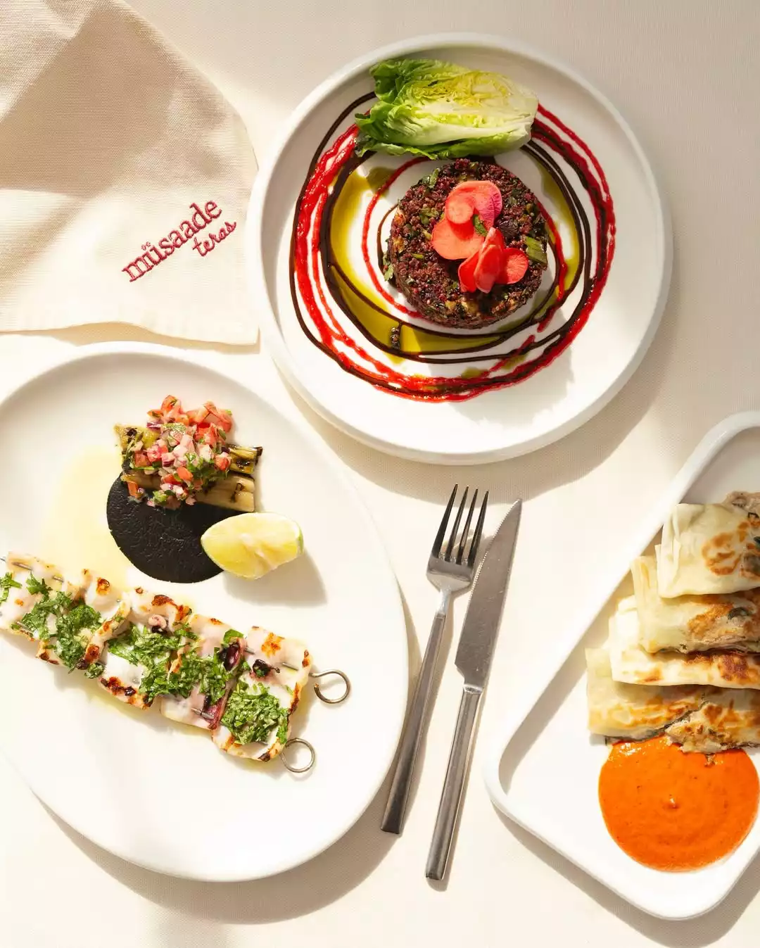 Şefin favori meyhanesi Müsaade Teras'ın modern meze masası. Parlak gün ışığında beyaz tabaklarda kırmızı soslu kinoa salatası, siyah soslu deniz ürünü şişi, börekler ve işlemeli mekan peçetesi zarif bir lezzet deneyimini