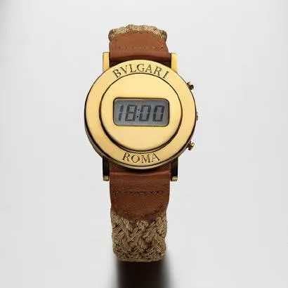 1975 BULGARI ROMA dijital sınırlı üretim altın saat, üzerinde BVLGARI ve ROMA yazan yuvarlak altın kasası, 18:00 gösteren dijital ekranı ve doğal örgülü kenevir ipi ile kahverengi deri karışımı kayışlarıyla beyaz bir zeminde duruyor.