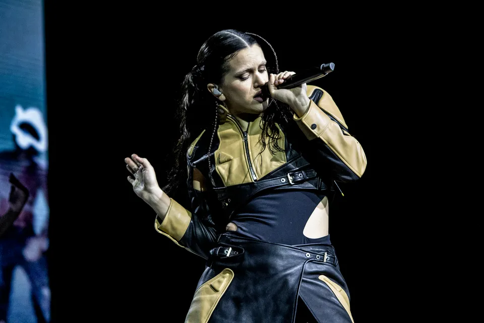Rosalia, siyah ve bej renkli, kemerli deri ceketi ile vücut hatlarını belli eden siyah kıyafeti içinde, gözleri kapalı ve mikrofonu sıkıca tutarak sahnede tutkuyla performans sergiliyor ve müzik dinleyicilerini büyüleyen güçlü sesiyle şarkı söylüyor.