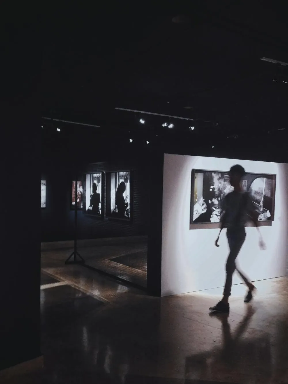 Modern bir sanat galerisinin loş atmosferinde, duvardaki spot ışıklarıyla aydınlatılmış, insanlar figürlerini gösteren siyah beyaz birçok sanat eserinin önünden bulanık bir şekilde hızla geçen bir ziyaretçi görülüyor; yansıyan ışıklar zeminde parlıyor.