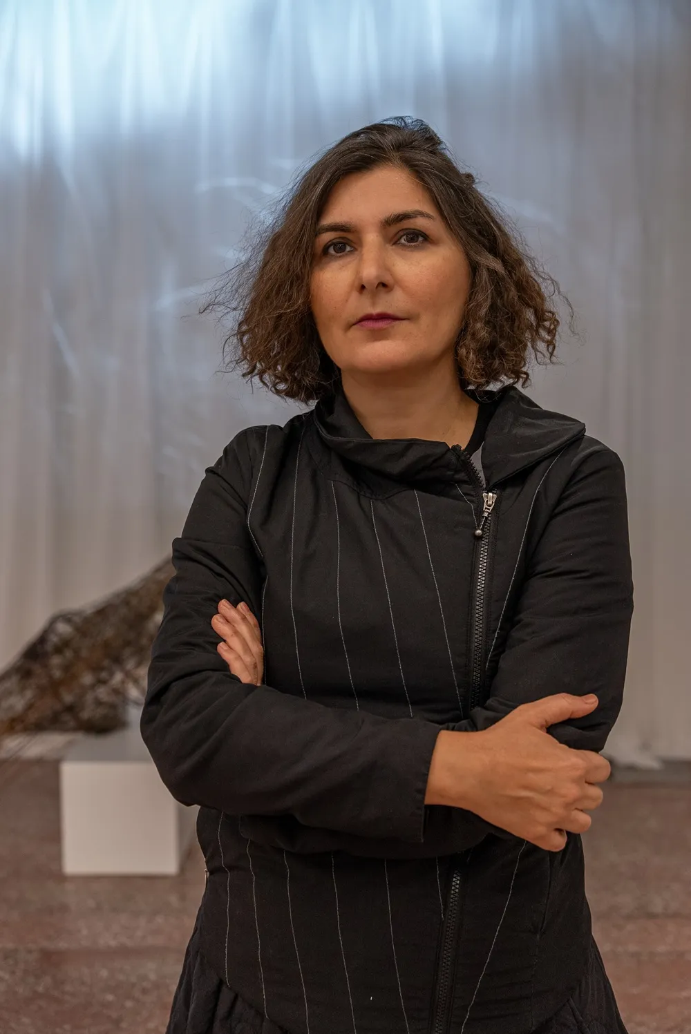 Şaqayeq Arabi, sanatçıya özgü duruşuyla, dalgalı kahverengi saçları ve çizgili siyah ceketiyle kameraya doğrudan bakarken, arkasında modern bir sanat eserinin ve beyaz bir kaidenin seçilebildiği bir sergi alanında kol kavuşturmuş poz veriyor.