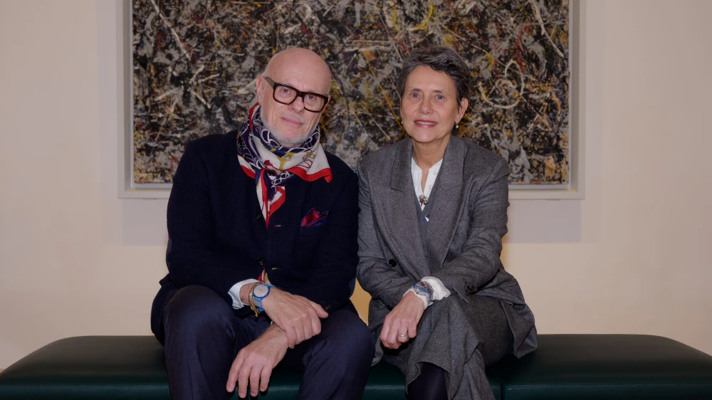 Swatch ve Guggenheim iş birliği bağlamında, koyu yeşil bir bankta yan yana oturan, arkalarında soyut bir tablo bulunan Guggenheim Müzesi Müdürü Karole Vail ve Swatch Kreatif Direktörü Carlo Giordanetti objektife gülümsüyor.