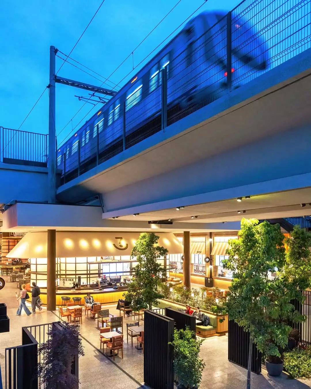 Oggusto Terminal Kadıköy Rehberi: Lezzet, Alışveriş ve Eğlence Durakları. Mavi akşam göğünde hızla geçen Marmaray treninin hemen altında hayat bulan canlı yaşam alanı. Ziyaretçiler, sıcak ışıklarla aydınlatılan 7DE7 lezzet durağında, yeşillikler içindeki masalarda oturarak sosyalleşiyor.