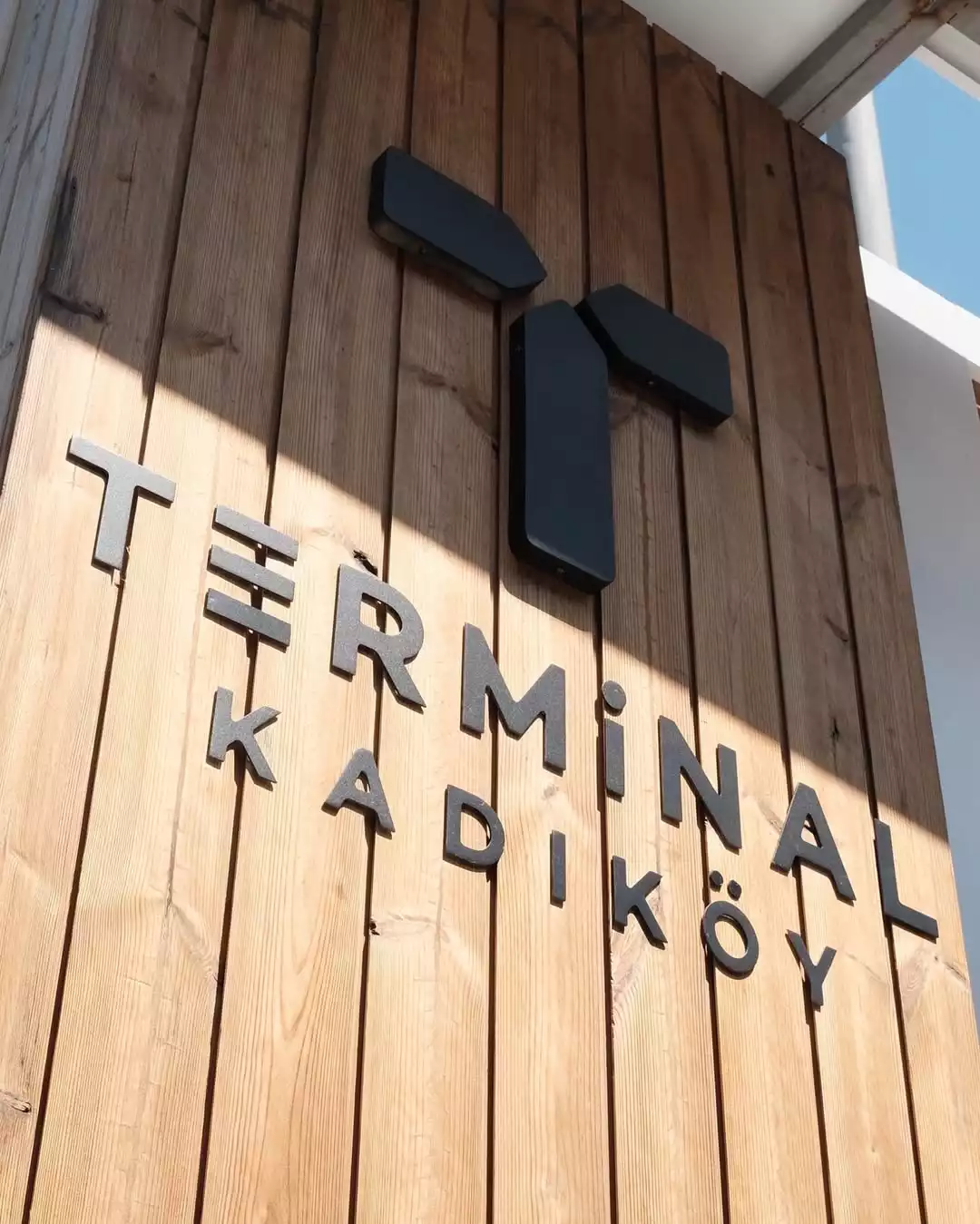 Oggusto Terminal Kadıköy Rehberi: Lezzet, Alışveriş ve Eğlence Durakları. Dikey ahşap paneller üzerinde siyah 3B harflerle vurgulanan Terminal Kadıköy tabelası ve parçalı T logosu, şehrin yeni buluşma noktasının dinamik kimliğini yansıtan keskin güneş gölgeleri altında sergileniyor.