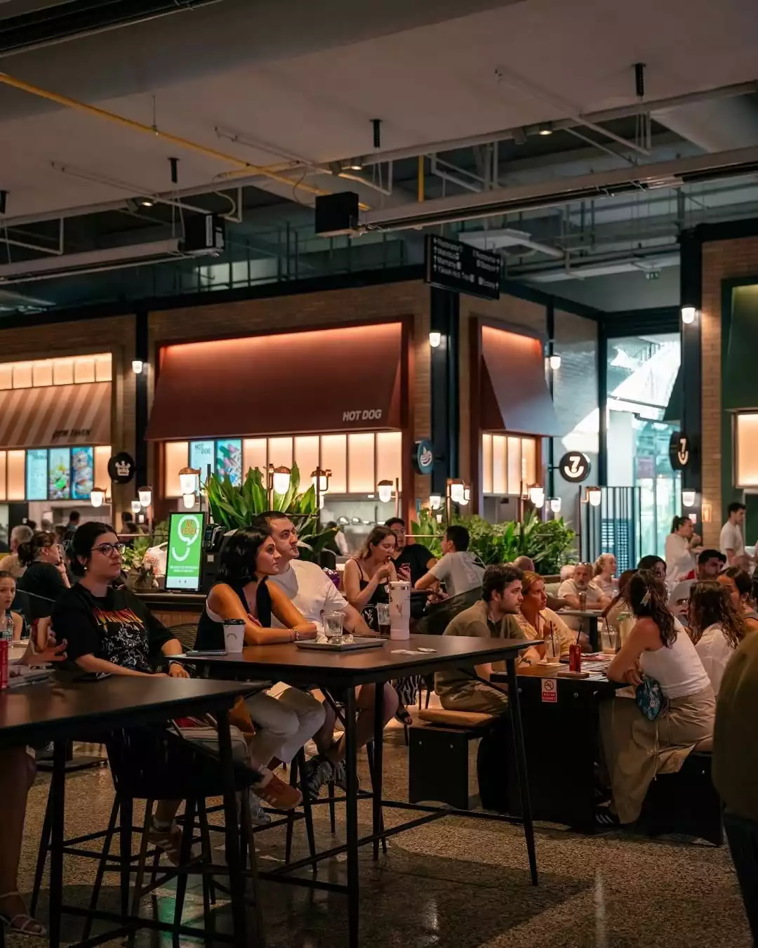 Oggusto Terminal Kadıköy Rehberi: Lezzet, Alışveriş ve Eğlence Durakları. Terminal Kadıköy'ün geniş 7DE7 yeme-içme alanında yüksek masalarda oturup sohbet eden ve yemek yiyen ziyaretçiler. Arka planda Hot Dog standı, bitkiler ve sıcak aydınlatmalarla modern endüstriyel gastronomi atmosferi.