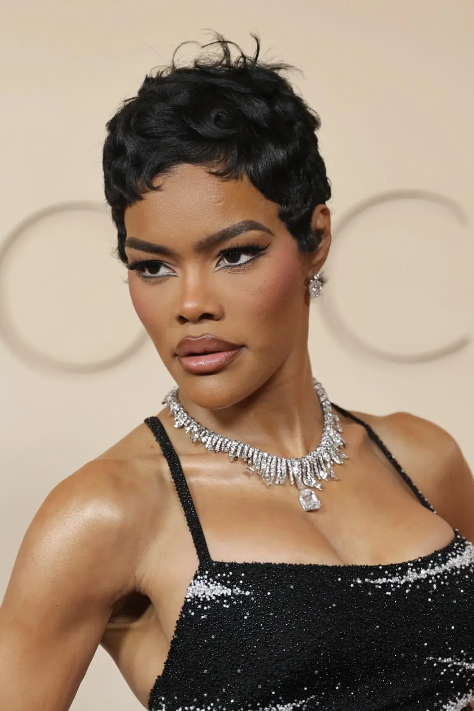 Teyana Taylor, kısa dalgalı siyah saçları, çekik eyeliner ile vurgulanmış gözleri, parlak dudakları ve ışıltılı siyah payetli, ince askılı elbisesinin üzerine taktığı göz alıcı elmas kolyesi ve küpeleriyle bir kırmızı halı etkinliğinde zarif ve etkileyici bir şekilde poz veriyor.