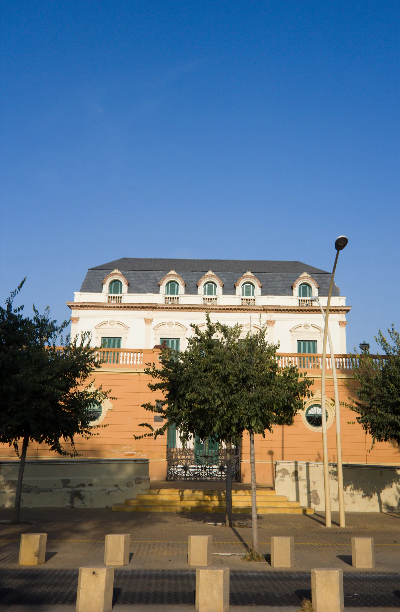 Sevilla'daki Alameda bölgesinde, koyu gri mansard çatılı, beyaz üst cephesi kemerli pencerelerle ve balkonu korkuluklarla süslü, alt kısmı turuncu renkte olan gösterişli bir bina, önündeki yeşil ağaçlar, modern bir sokak lambası ve beton bloklarla çevrili kaldırım ile mavi gökyüzü altında duruyor.
