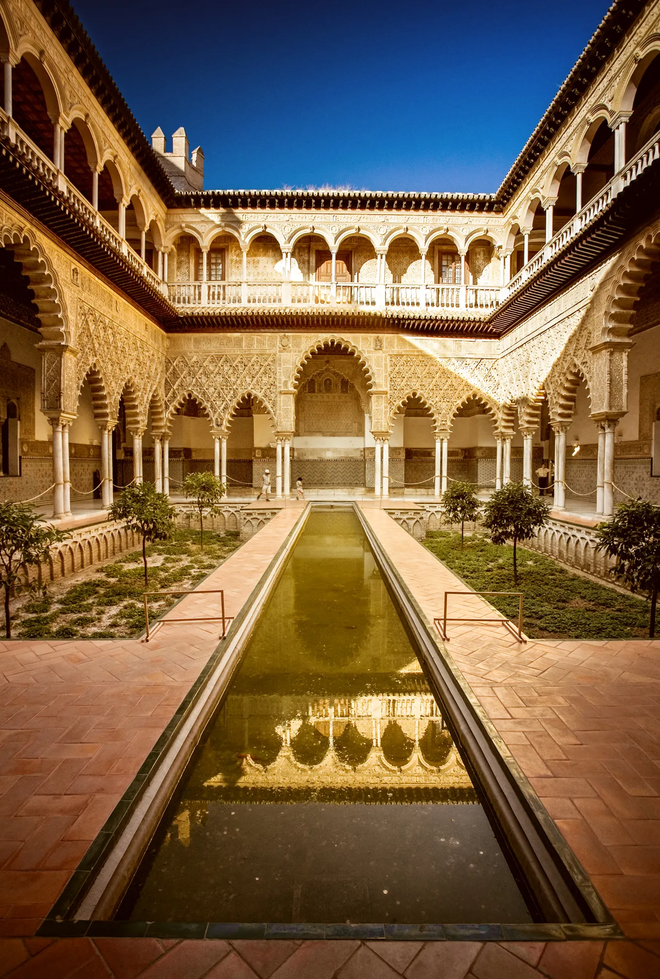 Mavi gökyüzünün altında, Sevilla Alcázar'ının Mudejar mimarisini yansıtan detaylı oymalı kemerler ve sütunlarla çevrili geniş avluda, ortadaki uzun, durgun su havuzu etrafındaki yeşil bahçeleri ve küçük ağaçları yansıtırken, güneş ışıkları sarayın zengin dokusunu aydınlatıyor.