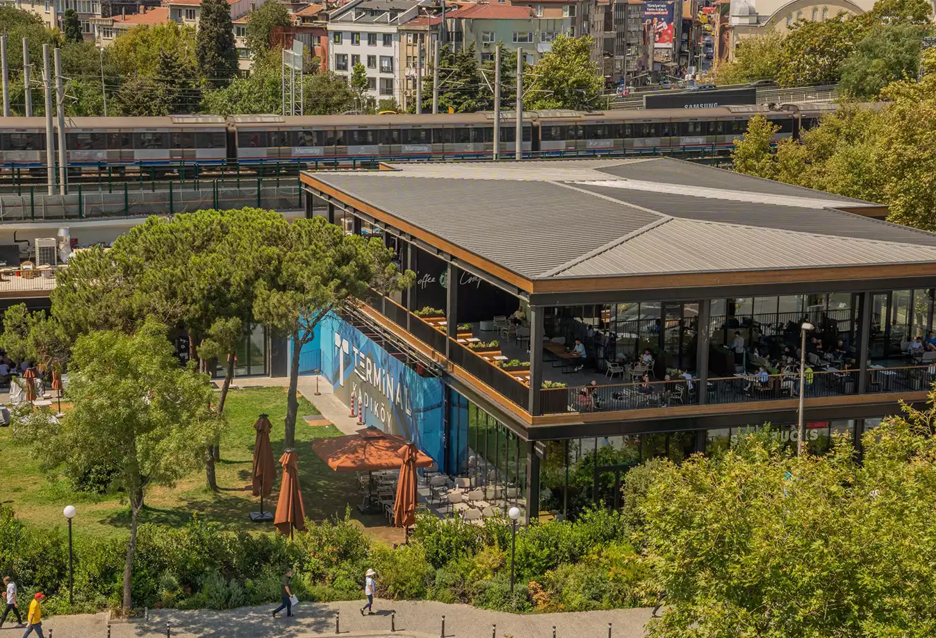 Oggusto - Terminal Kadıköy Rehberi: Lezzet, Alışveriş ve Eğlence Durakları. Arka planda geçen Marmaray treninin hemen yanında, yeşil alanlarla çevrili iki katlı cam mimarili Terminal Kadıköy binası. Ziyaretçiler yeni yaşam alanının açık hava teraslarında sosyalleşiyor.