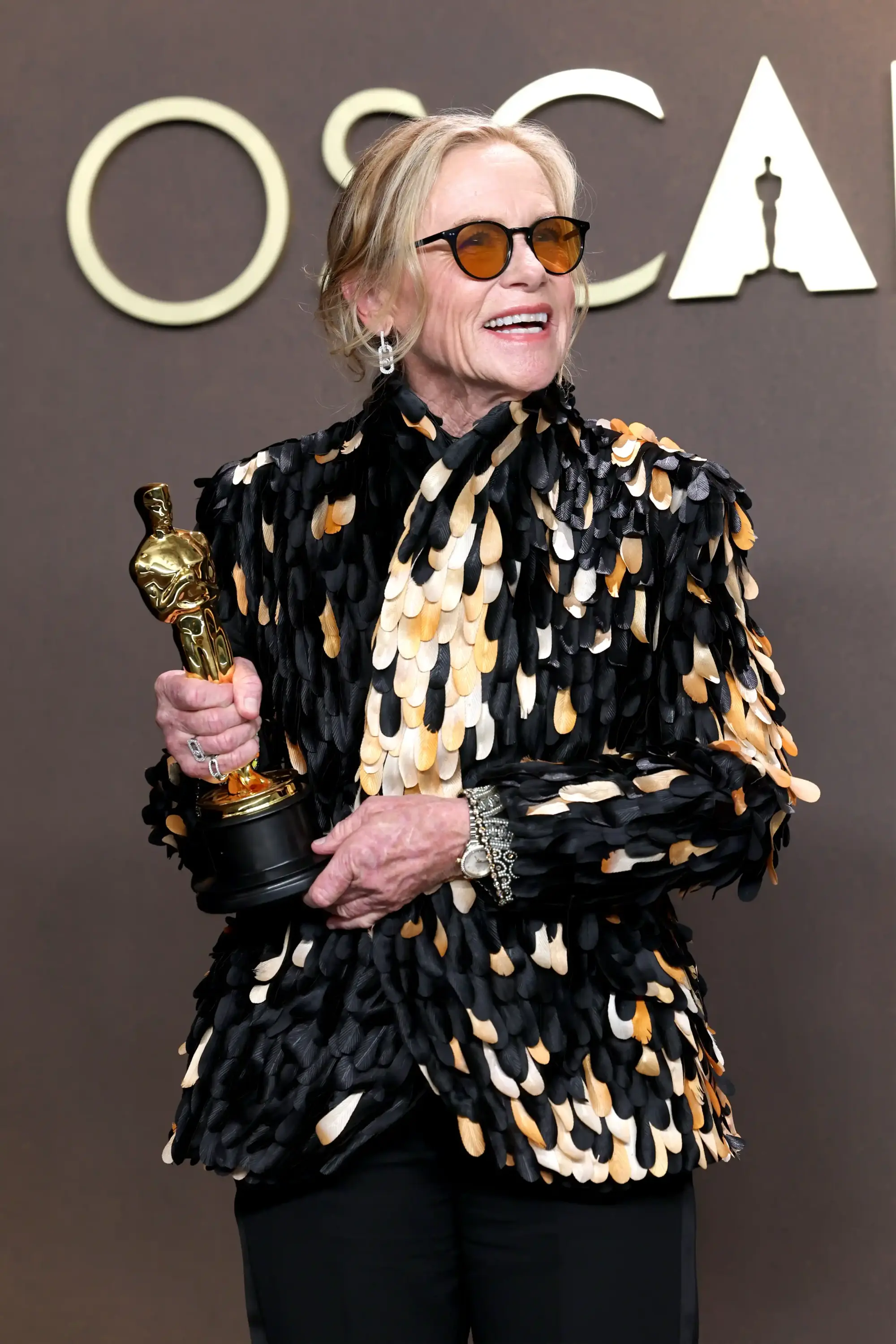 Amy Madigan, siyah, beyaz ve turuncu tonlarda tüy/pul detaylı bir ceket giymiş, turuncu camlı güneş gözlükleriyle geniş bir gülümsemeyle Oscar heykelciğini tutarken, arka planda OSCAR logosu beliriyor.