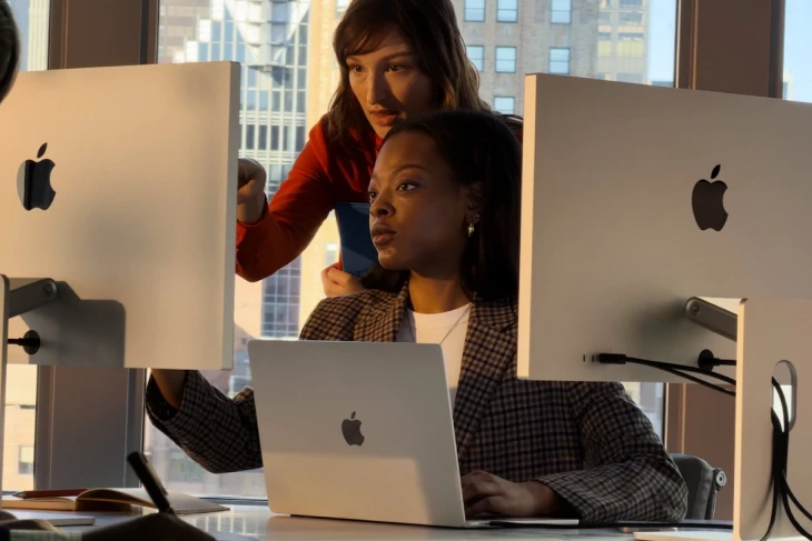 İki profesyonel kadın, modern bir ofis ortamında, şehir manzarası önünde Apple MacBook Pro ve Studio Display ekranlarını kullanarak işbirliği içinde çalışırken, bu görüntü yeni nesil Apple M5 çip ailesiyle güçlendirilmiş MacBook Pro'ların Yapay Zeka Performansını ve profesyonel iş akışlarındaki verimliliğini vurguluyor.