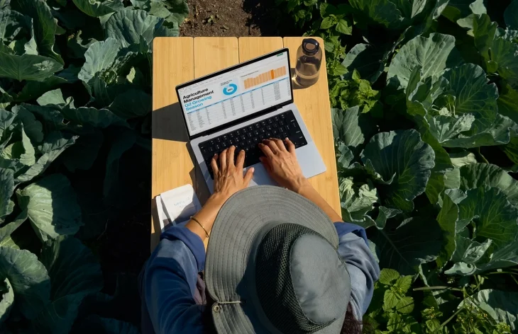 Geniş kenarlı şapka takan bir kişinin, yemyeşil bitkilerle dolu bir tarlanın ortasında ahşap bir masa üzerinde duran Apple MacBook Pro dizüstü bilgisayarında tarım yönetimi verilerini gösteren karmaşık bir elektronik tablo üzerinde çalıştığı yukarıdan çekilmiş bir görüntü, Apple M5 işlemcili MacBook modellerinin yapay zeka performansı ile mobil profesyonel iş akışlarını her yerde mümkün kıldığını vurguluyor.