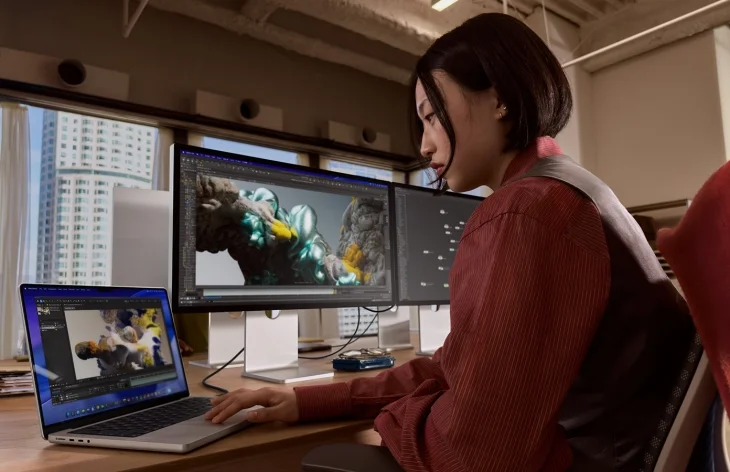 Kırmızı bir gömlek giymiş genç bir kadın, Apple'ın M5 Pro veya M5 Max çipli MacBook Pro'sunu ve iki büyük ekranı kullanarak, yapay zekâ destekli video kurgu veya 3D modelleme yazılımlarında karmaşık projeler üzerinde yoğunlaşıyor.