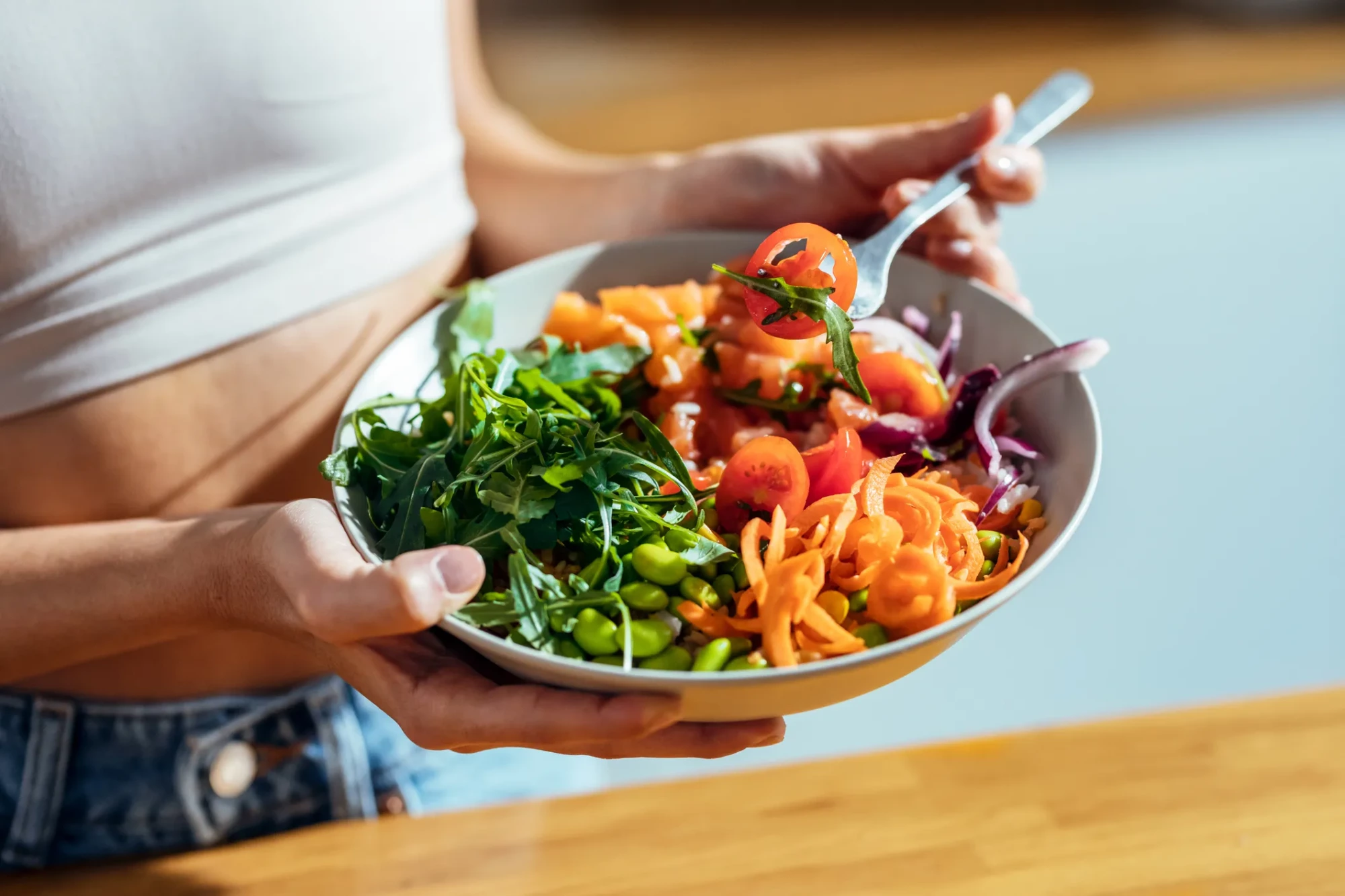 Beyaz bir atlet ve kot pantolon giyen bir kişi, taze roka, kiraz domates, rendelenmiş havuç, edamame ve kırmızı soğan içeren rengarenk, sağlıklı bir salata kasesini tutuyor, çatalında bir parça domates ve roka var, bu görüntü Başak burcu için önerilen Nisan ayı detoks enerjisi ve sağlık temasına uygun beslenmeyi vurguluyor.