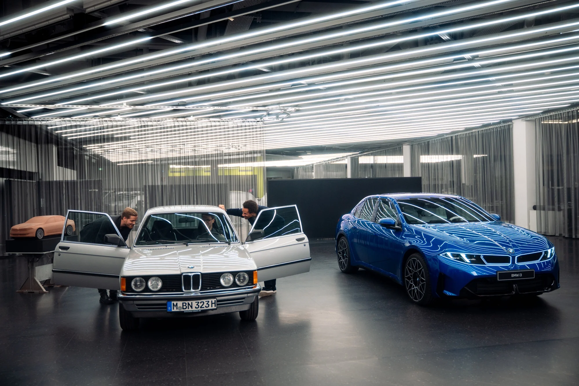 Münih'teki BMW tasarım ofisinde, iki kişinin kapıları açık gümüş renkli klasik bir BMW 3 Serisi otomobilin içini incelediği, sağda ise modern tasarım hatlarına sahip parlak mavi bir BMW'nin durduğu ve arka planda bir araç kil modelinin de sergilendiği, geleceğin ve geçmişin tasarım felsefesini buluşturan geniş bir stüdyo ortamı.