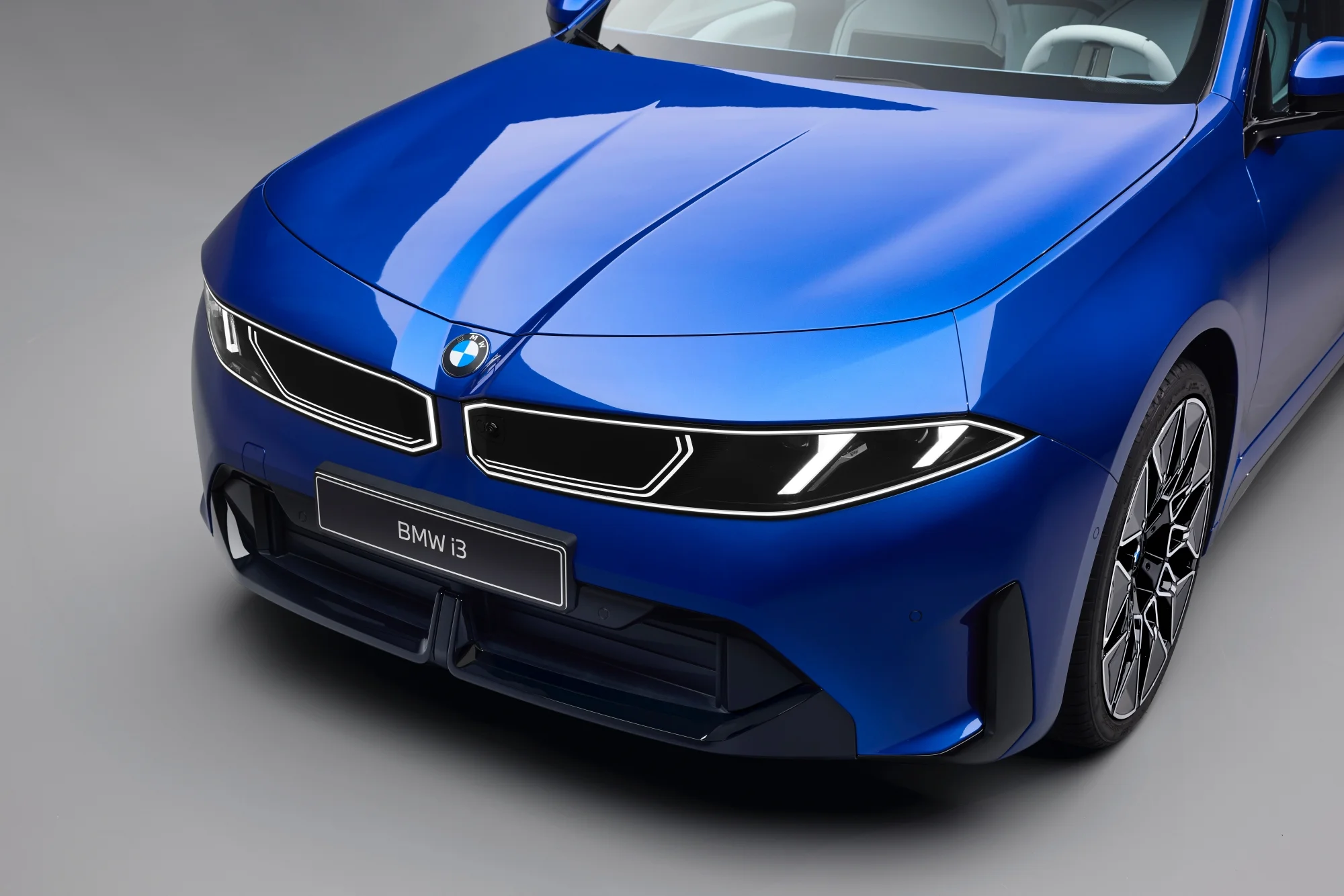 Parlak mavi renkteki yeni BMW i3'ün ön görünümü, marka logosu, modern yorumlu böbrek ızgaraları ve keskin LED farlarıyla dikkat çekerken, özellikle geleceğe yönelik Neue Klasse tasarım dilini sergiliyor.