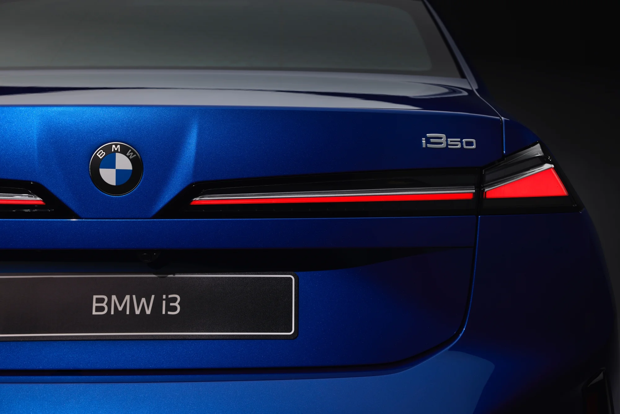 Parlak mavi renkteki BMW i350 elektrikli otomobilin arkasını vurgulayan yakın çekim, bagaj kapağının merkezinde klasik BMW logosunu, sağda gümüş renkli i350 model ismini ve alt kısımda BMW i3 yazısı bulunan siyah plakalık çerçevesiyle uzanan zarif kırmızı LED stop lambaları detaylarını ortaya koyuyor.