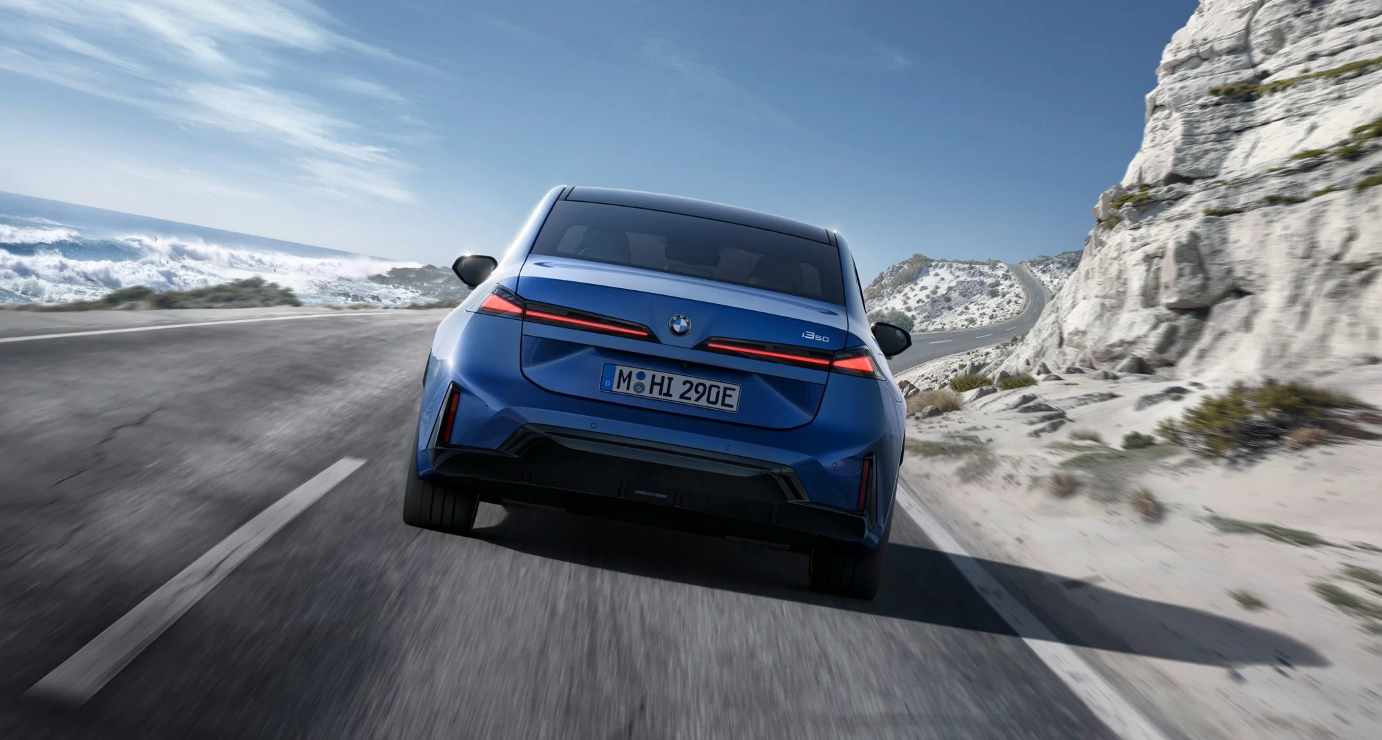 Mavi renkli bir BMW i350, modern arka tasarımı ve keskin kırmızı stop lambalarıyla, solunda büyük dalgaların olduğu masmavi okyanus, sağında ise beyaz kayalıklar ve kumlu tepelerin uzandığı kıvrımlı bir yolda hızlıca ilerlerken görüntülendi. Bu, BMW'nin geleceğe dönük Neue Klasse lansman vizyonunu yansıtan dinamik bir çekim.