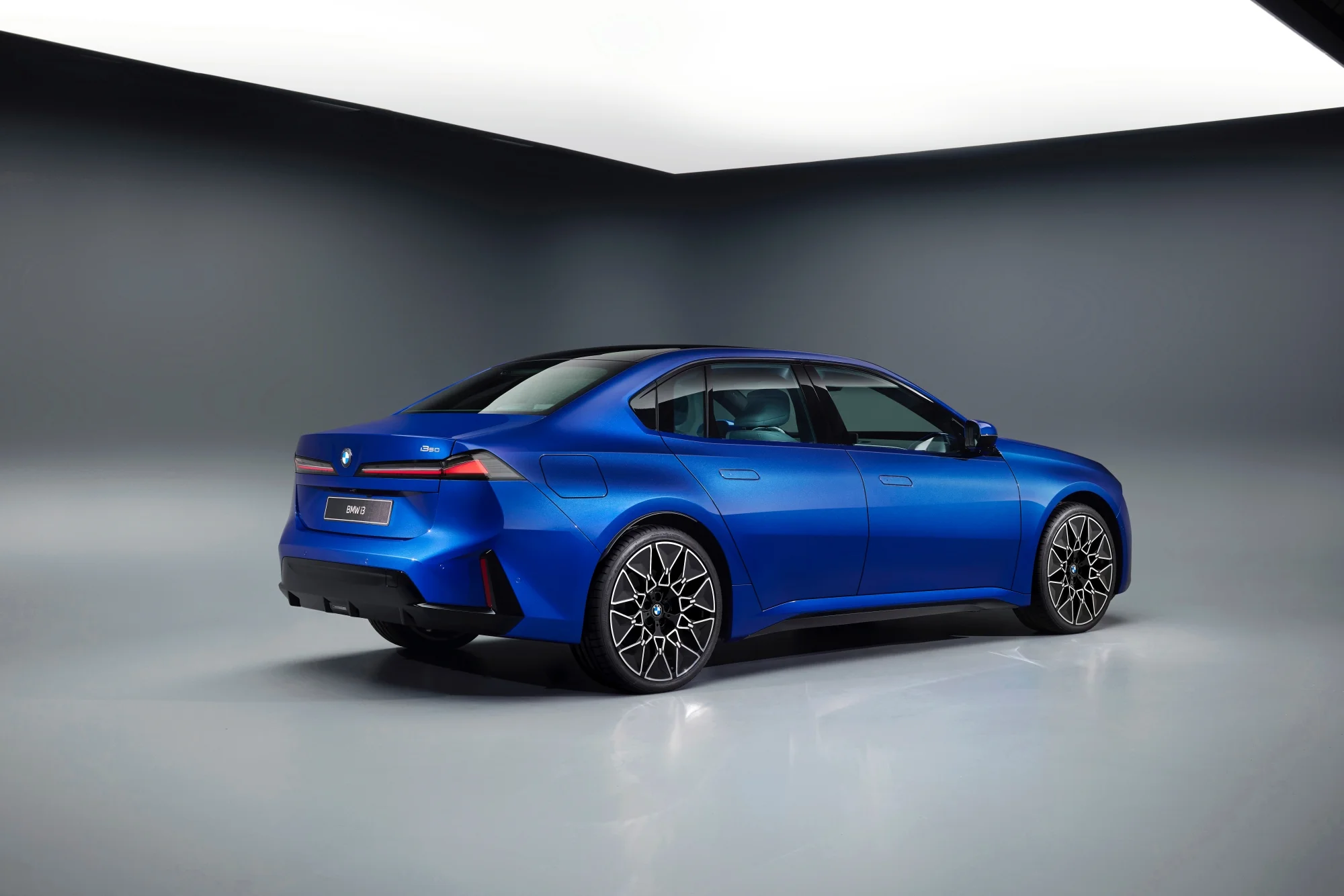 Mavi renkteki BMW Neue Klasse i3 sedan, stüdyo ortamında arka üç çeyrek açıdan sergilenirken modern ve aerodinamik arka tasarımını, dikkat çekici LED stop lambalarını, siyah tavanını ve özgün desenli jantlarını gözler önüne seriyor.