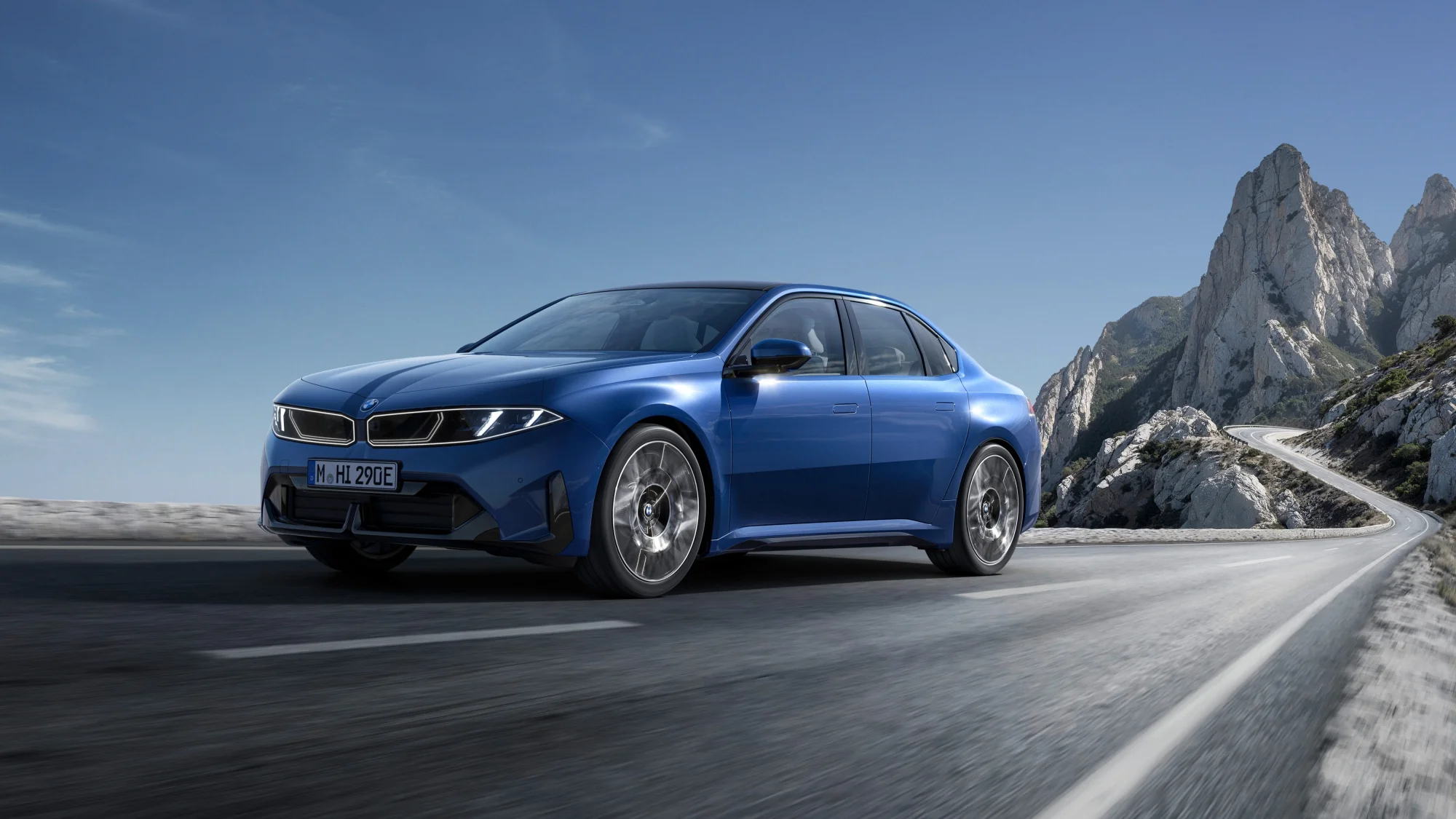 Mavi renkli modern bir BMW Neue Klasse sedan otomobil, güneşli bir günde, tekerleklerinde hafif bir hareket bulanıklığıyla virajlı bir dağ yolunda ilerliyor; arka planda heybetli, gri tonlu kayalık zirveler ve açık mavi bir gökyüzü dikkat çekiyor.