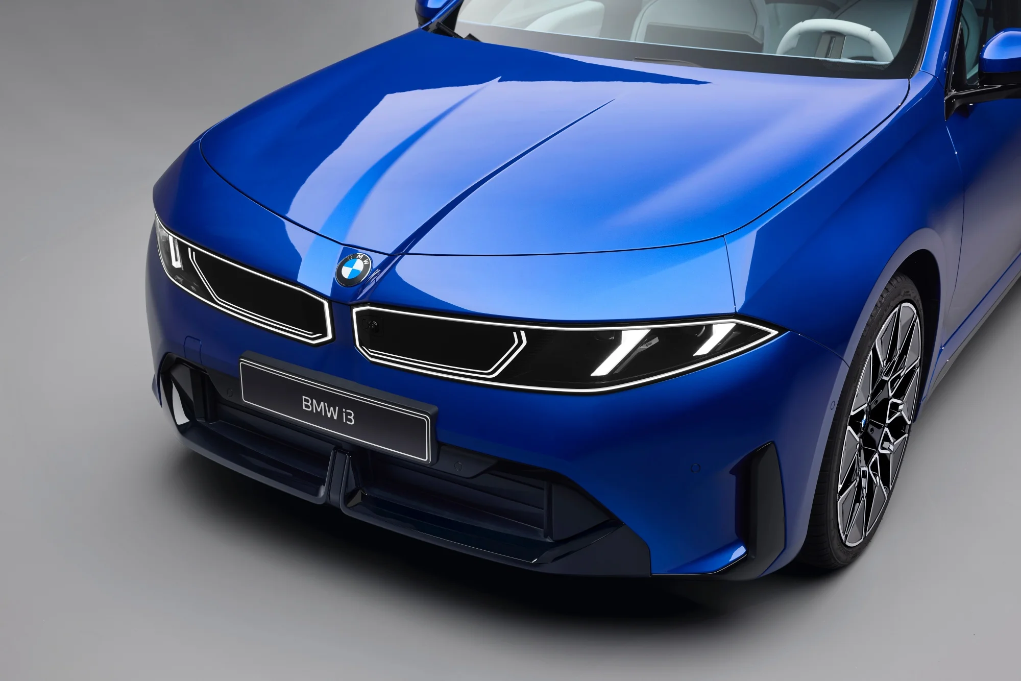 Parlak mavi renkteki yeni nesil BMW i3'ün ön sol açıyla çekilmiş yakın plan fotoğrafında, aracın belirgin LED aydınlatmalı modern böbrek ızgarası, keskin hatlı farları ve akıcı dış tasarım çizgileri vurgulanıyor.