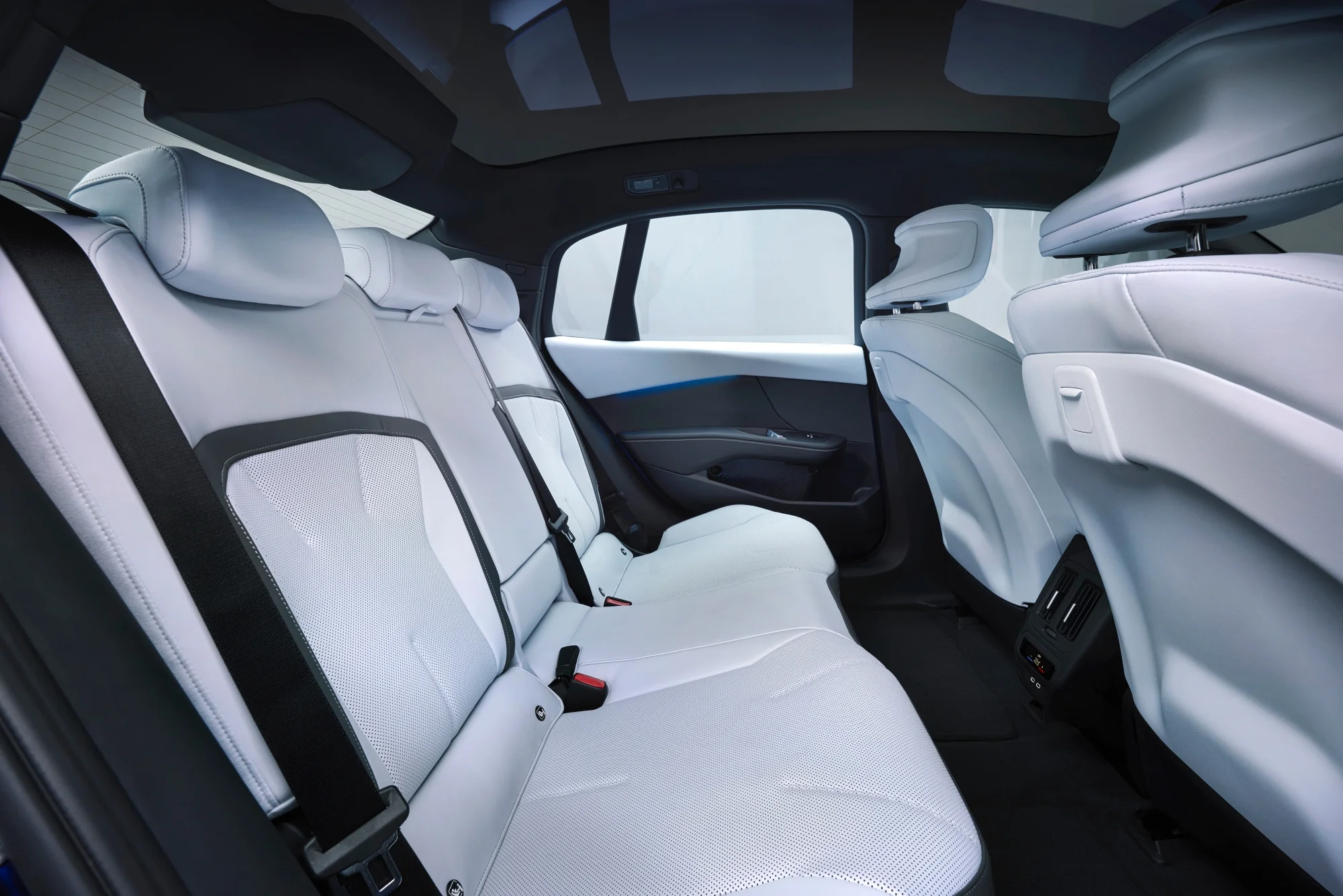 BMW Neue Klasse i3'ün modern iç mekan tasarımını sergileyen bu görüntü, açık renk delikli deri kaplı ferah arka koltukları, koyu tavan ve kapı panelindeki zarif mavi ambiyans aydınlatmasıyla dikkat çekerken, arka orta konsoldaki dijital ekran ve USB portları işlevselliği vurguluyor.