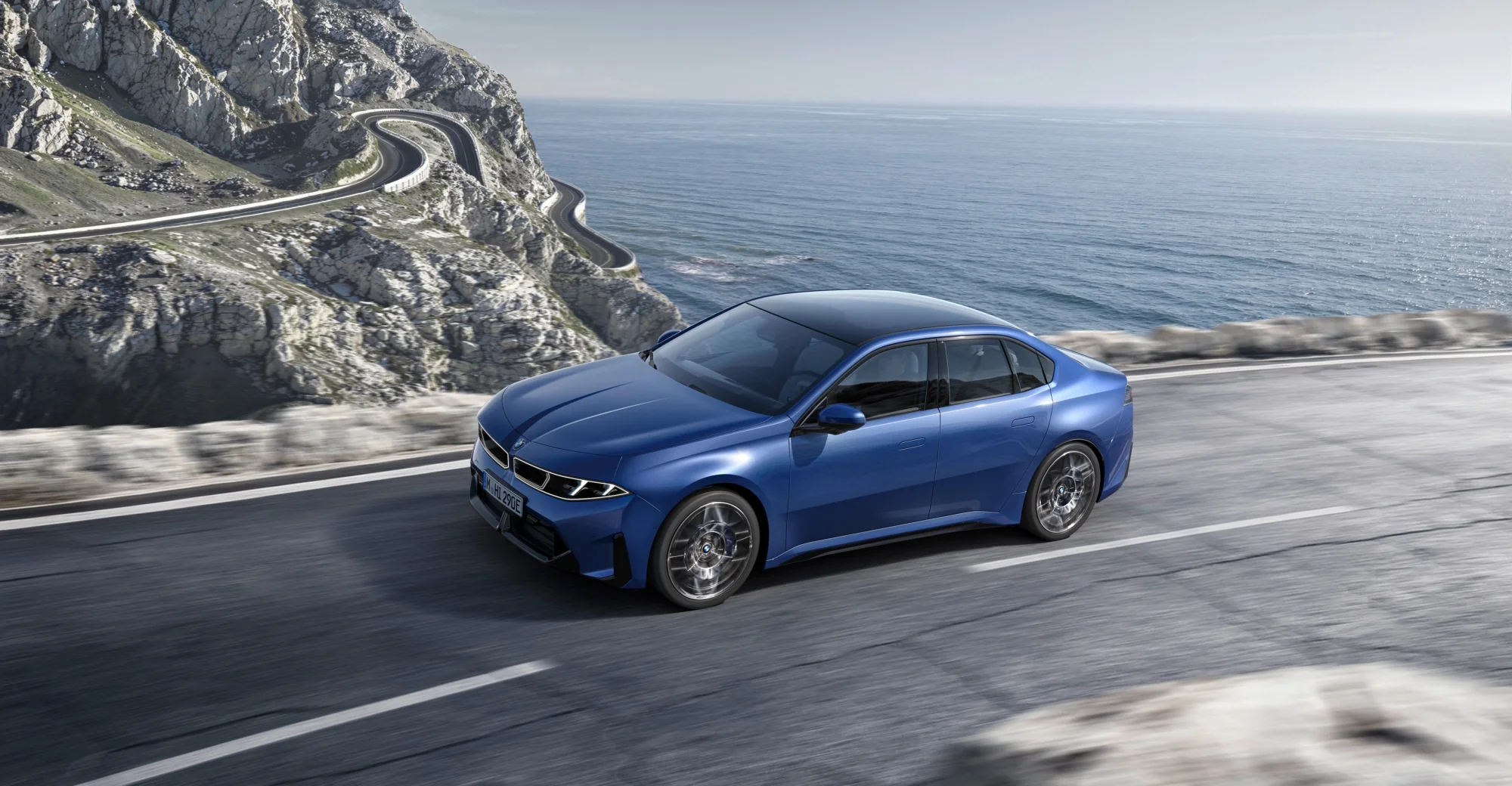 Masmavi bir BMW Neue Klasse i3, dağlık ve kıvrımlı bir sahil yolunda hızla ilerlerken, solunda sarp kayalıklar ve sağında engin masmavi denizle çevrili nefes kesici bir manzarada dinamik bir şekilde görüntüleniyor.