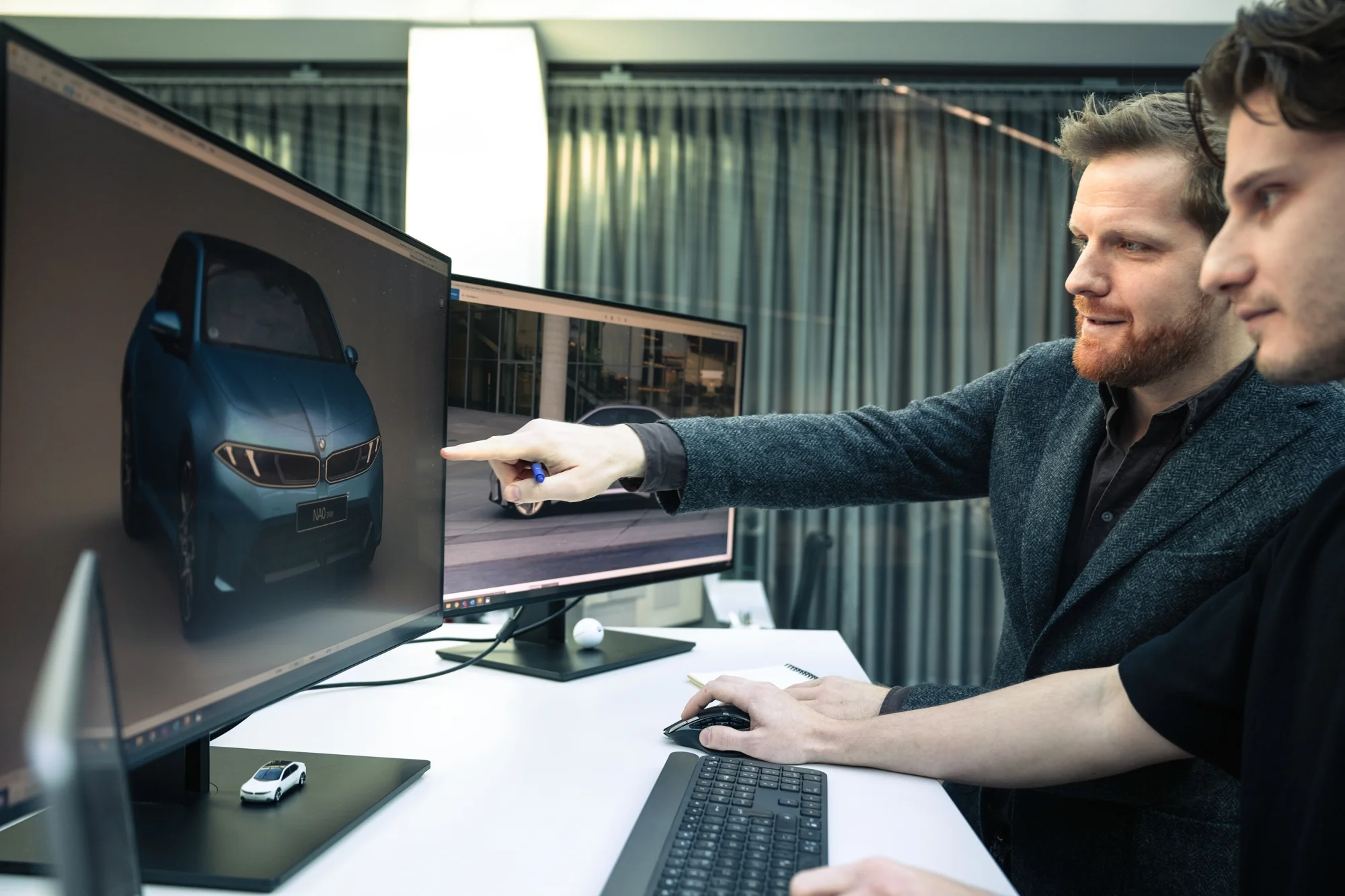 Bir ofis ortamında iki tasarımcı, geleceğin BMW Neue Klasse araçlarından birinin dijital tasarımını büyük bir ekranda detaylı bir şekilde inceliyor; sakallı olan parmağıyla aracın ön farlarını işaret ederken, masadaki minyatür model araba bu yoğun tasarım süreci çalışmasının bir parçası olarak görülüyor.