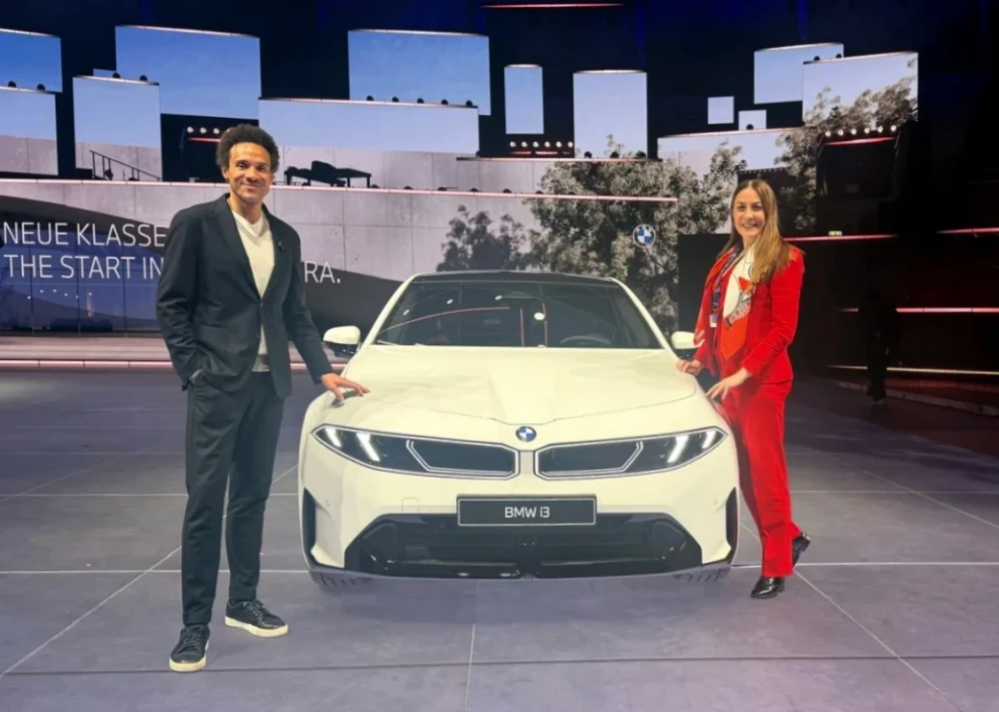 BMW Tasarım Başkanı Oliver Heilmer ve Oggusto'dan Özlem Gusar, beyaz BMW i3 modelinin önünde gülümsüyorken, şık kıyafetleriyle aracın kaputuna ellerini koyarak bir röportaj kapsamında poz veriyorlar.