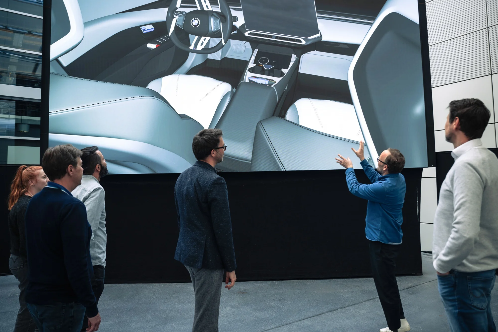 Münih'teki bir BMW tasarım ofisinde, bir grup tasarımcı ve mühendis, büyük bir ekranda gösterilen yeni bir BMW modelinin modern iç tasarım detaylarını tartışırken, sunum yapan bir kişi elleriyle açıklama yapıyor.