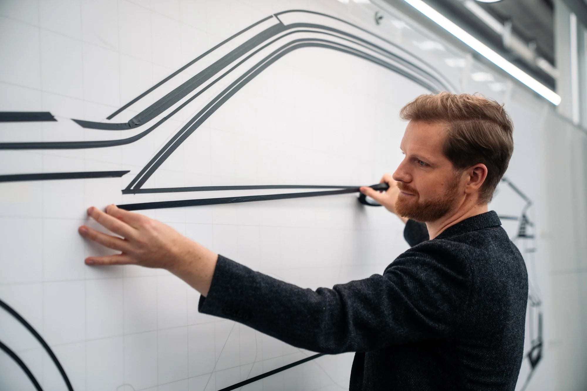 Sakallı bir tasarımcı, BMW tasarım ofisinde büyük bir panoya Neue Klasse elektrikli araçlarının gelecekteki çizgilerini oluşturmak için siyah bantla bir otomobilin yan profilini dikkatlice şekillendiriyor.