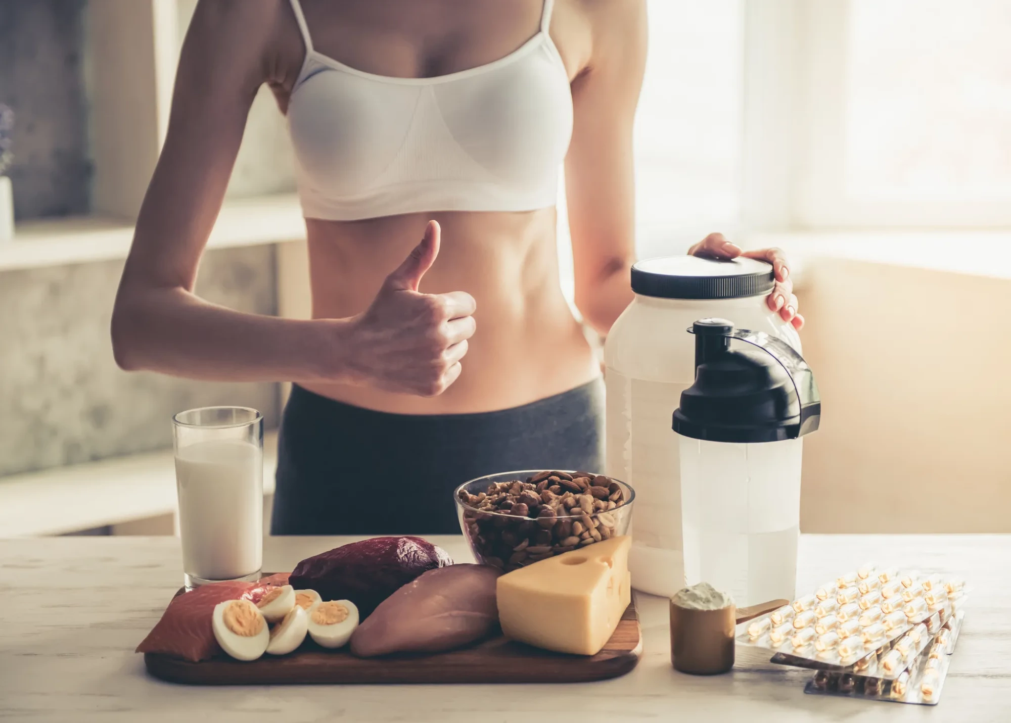 Beyaz spor sütyeni giymiş fit bir kadın, masada duran et, yumurta, peynir, kuruyemiş gibi protein kaynakları, süt, protein tozu ve vitamin desteklerinin önünde başparmağını yukarı kaldırarak Boğa burcunun Nisan 2026 sağlık tavsiyeleriyle uyumlu dengeli beslenmenin ve vitamin desteğinin önemine vurgu yapıyor.