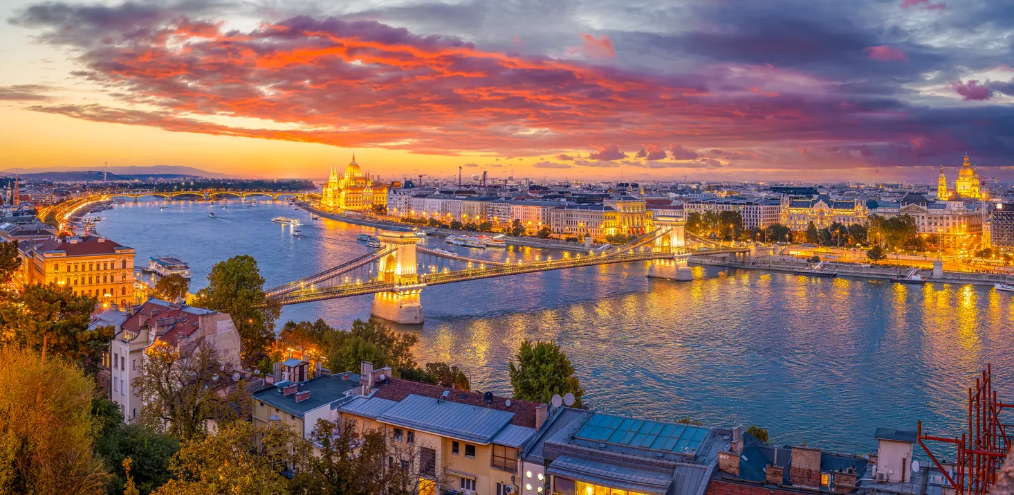 Gün batımında Budapeşte'nin büyüleyici panoramik görüntüsü, Tuna Nehri üzerinde ışıklandırılmış Zincirli Köprü ve Macaristan Parlamento Binası'nın altın rengi parıltısı ile bir seyahat destinasyonu olarak şehrin güzelliğini vurguluyor.