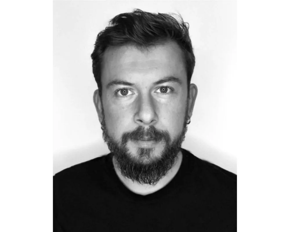 Siyah beyaz çekilmiş, hafif dalgalı koyu saçlı, sakallı ve kulaklarında küçük halka küpeler olan Burak Uzen'in kameraya doğrudan baktığı, koyu renk tişört giydiği ve Tiyatro Günü bağlamında anlam kazanan sade beyaz fonda bir portre fotoğrafı.