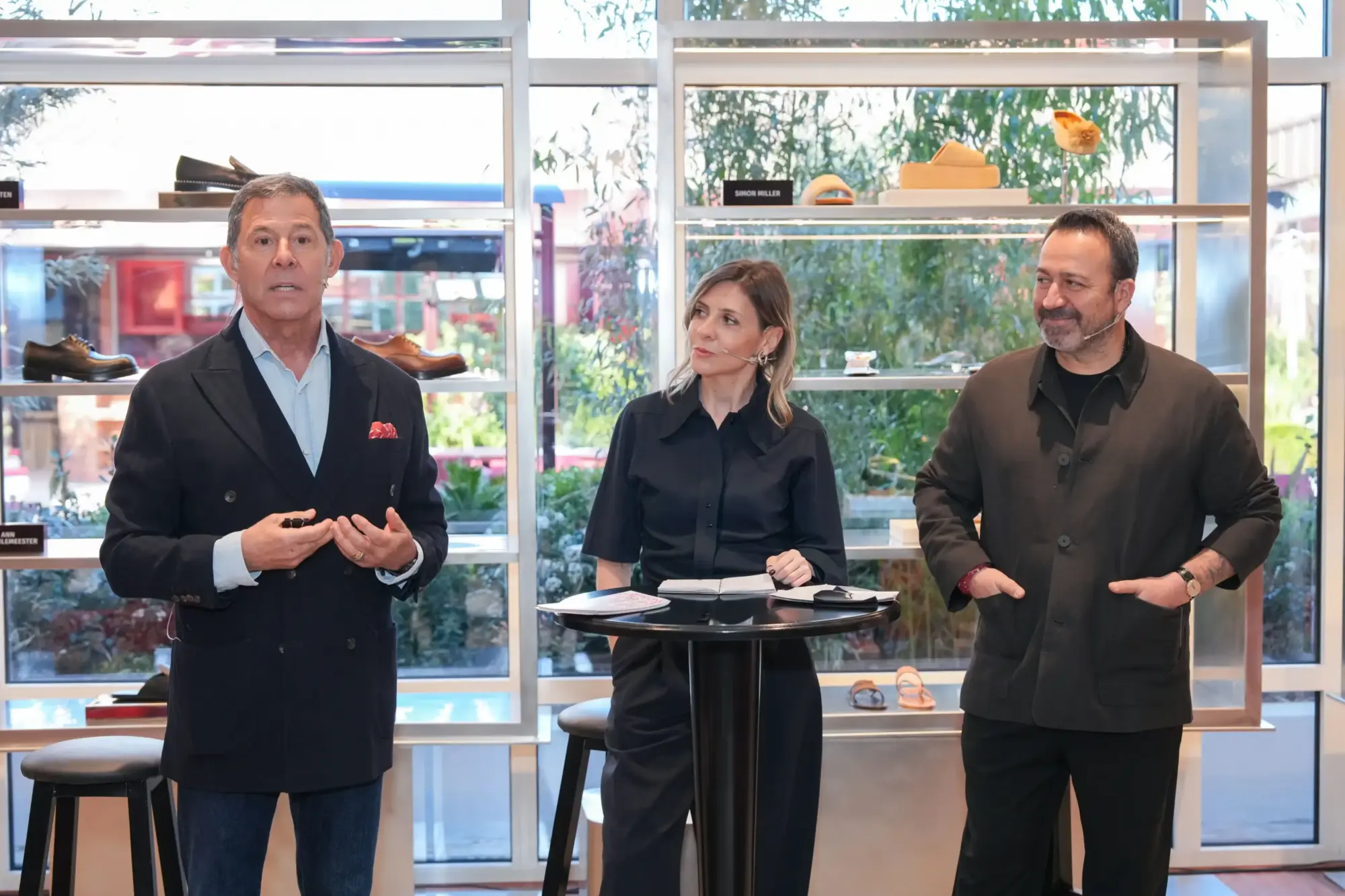 Cem Boyner, modern bir perakende alanında, arkasında sergilenen Ann Demeulemeester ve Simon Miller gibi markaların yeni tasarımları olan ayakkabılar eşliğinde, bir marka gelişimleri veya özel iş birliği etkinliğinde konuşma yaparken diğer katılımcılar onu dinliyor.