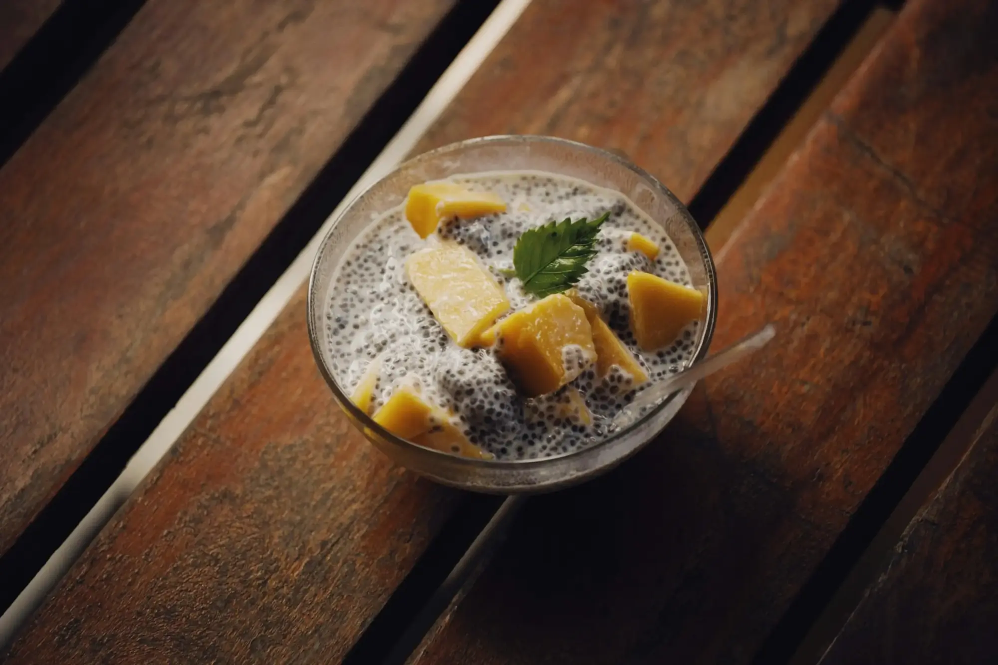 Koyu renk ahşap masa üzerinde, şişmiş chia tohumlarından yapılmış, üzerine canlı sarı mango küpleri ve taze yeşil bir nane yaprağı eklenmiş, sağlıklı ve besleyici bir chia pudingi kasesi görülüyor; bu sunum chia tohumunun çeşitli faydalarından yararlanmak için ideal bir örnektir.