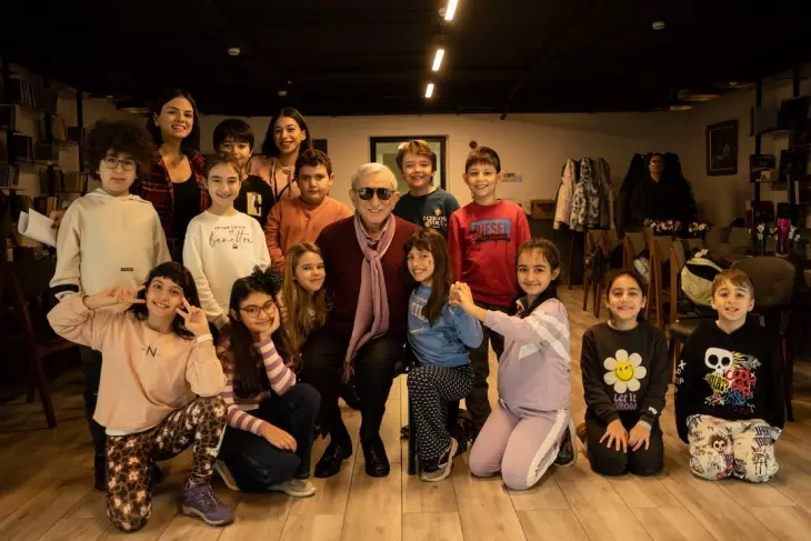 Çocuklar için yaratıcı drama ve tiyatro kursları kapsamında bir araya gelen çeşitli yaşlardaki öğrenciler, liderleri olduğu düşünülen tecrübeli bir sanatçı ve genç eğitmenlerle birlikte iç mekanda neşeyle poz veriyor, eğitim sürecindeki mutluluklarını yansıtıyor.