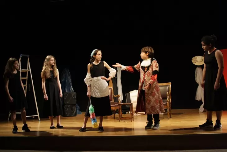 Bu görüntü, çocukların yaratıcı drama veya tiyatro kurslarında sahne becerilerini geliştirirken eğlenerek farklı rollere büründüğünü gösteriyor; sahnedeki beş çocuktan biri hizmetçi kostümüyle renkli bir toz püskülü tutarken, diğeri eski dönem soylu kıyafetleriyle ona bir şeyler anlatırcasına kolunu uzatıyor, bu da onların hayal güçlerinin ve ifade yeteneklerinin gelişimine vurgu yapıyor.