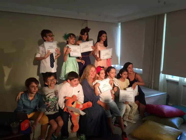 Kapalı alanda çekilen bu fotoğrafta, tiyatro eğitimi almış gibi duran, bazıları bıyık takmış ve süslü saç aksesuarları olan çocuklarla birlikte, tamamladıkları yaratıcı drama veya tiyatro kurslarının sertifikalarını gururla tutan yetişkinler ve çocuklar neşeyle poz veriyor.