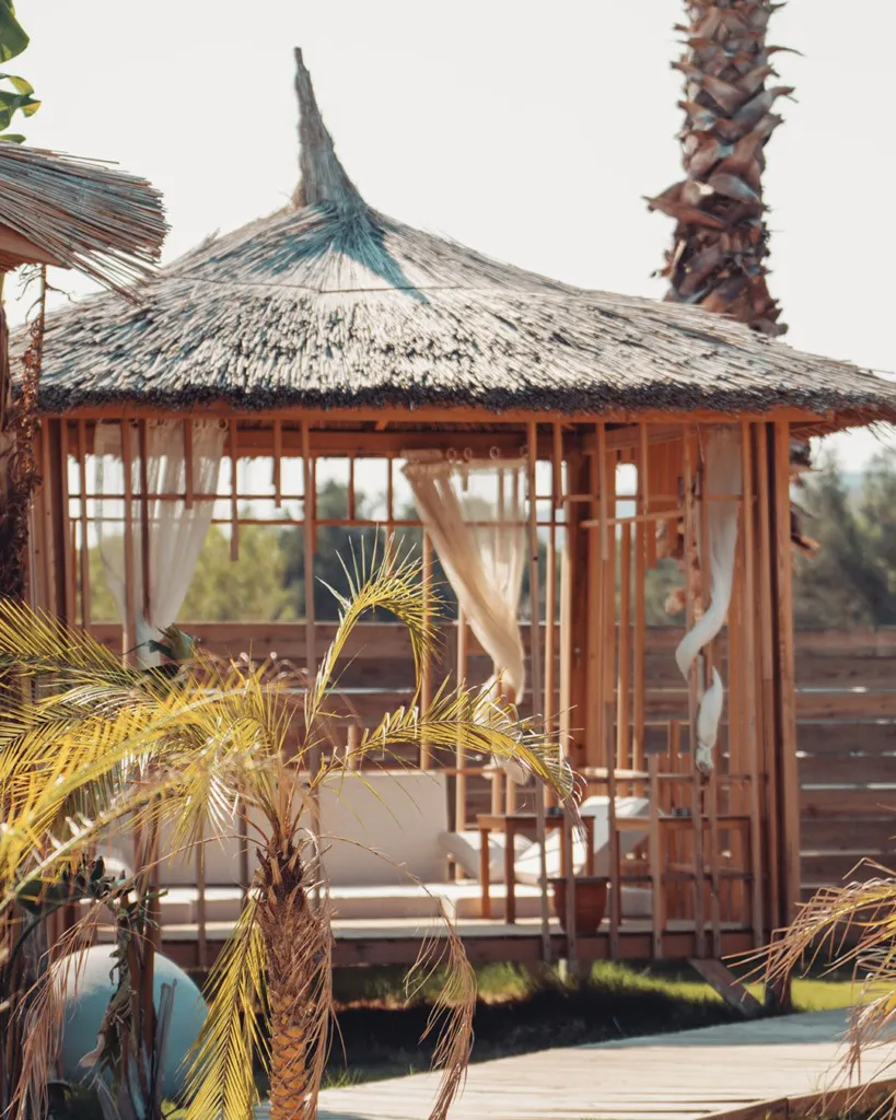 Cunda Ajlan Beach'teki hasır çatılı ve bembeyaz tüllerle çevrili bu ahşap çardak, içerideki konforlu beyaz minderli oturma alanıyla, etrafındaki palmiye ağaçları ve ahşap zeminle birlikte huzurlu bir dinlenme köşesi sunuyor.
