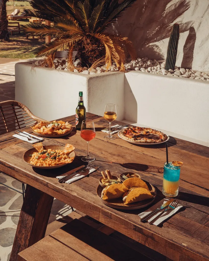 Cunda Ajlan Beach'te güneşli bir öğleden sonra, dış mekandaki ahşap bir masada taze pizzalar, kremalı makarnalar, çıtır takolar, kızarmış deniz ürünleri ve rengarenk içecekler iştah açıcı bir şekilde servis edilmiş, dinlendirici bir yaz tatili atmosferi sunuyor.