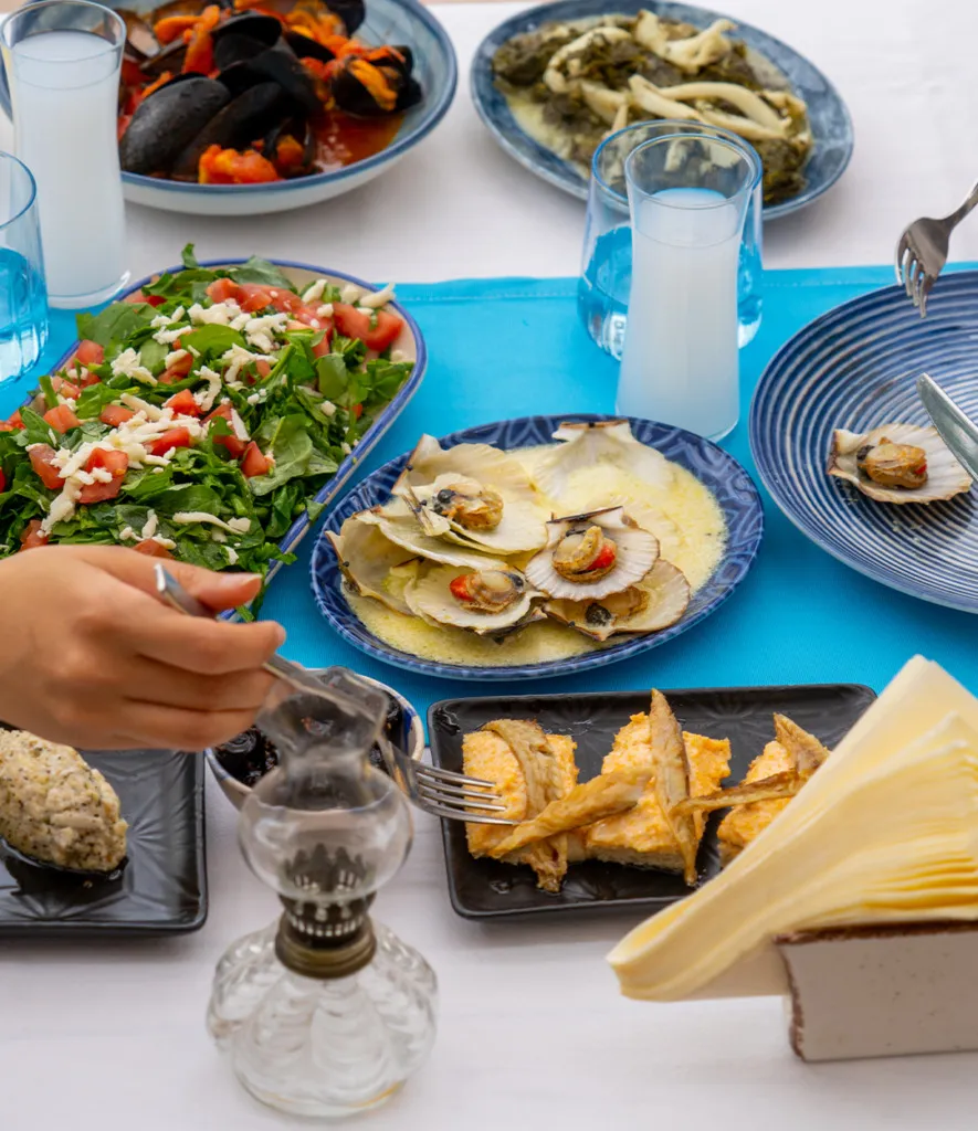 Cunda'da gezilecek yerler deneyimini tamamlayan, masada bol çeşitli deniz ürünleri, midye, tarak, taze yeşil salata ve rakı kadehleriyle donatılmış, misafirlerin keyifle yemek yediği otantik bir Ege sofrası uzanıyor.