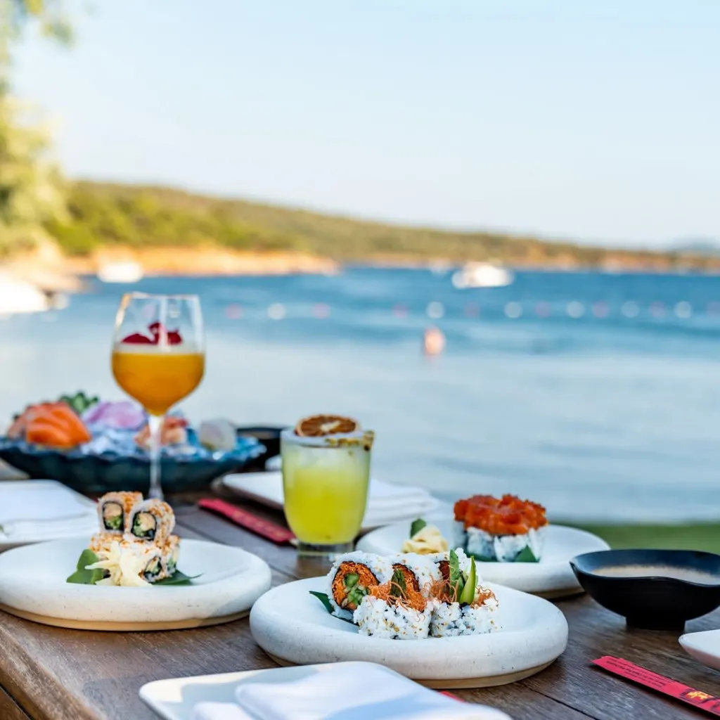 Çeşitli sushiler, iki renkli içecek ve sos kasesinin ahşap bir masada masmavi denize ve yemyeşil Cunda kıyılarına nazır keyifli bir yemek deneyimi sunulduğu huzurlu bir dış mekan restoranı.
