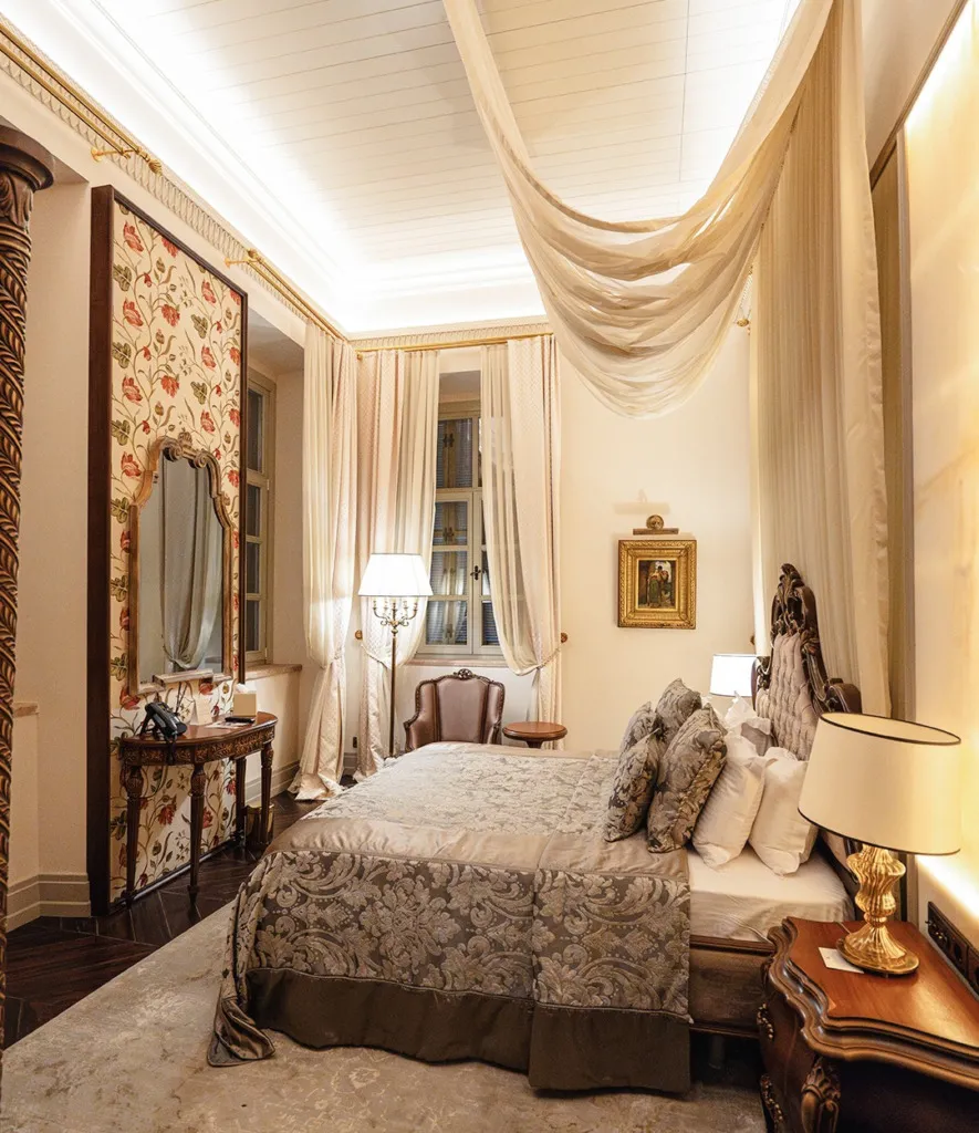 Zarif bir butik otel odasında, oymalı ahşap yatak başlığı, tavanından sarkan krem rengi şık bir tül perde, desenli yatak örtüsü ve yanındaki aynalı konsol masasıyla süslenmiş, Cunda'da konforlu bir konaklama için lüks ve geleneksel detayları bir araya getiren davetkar bir atmosfer.