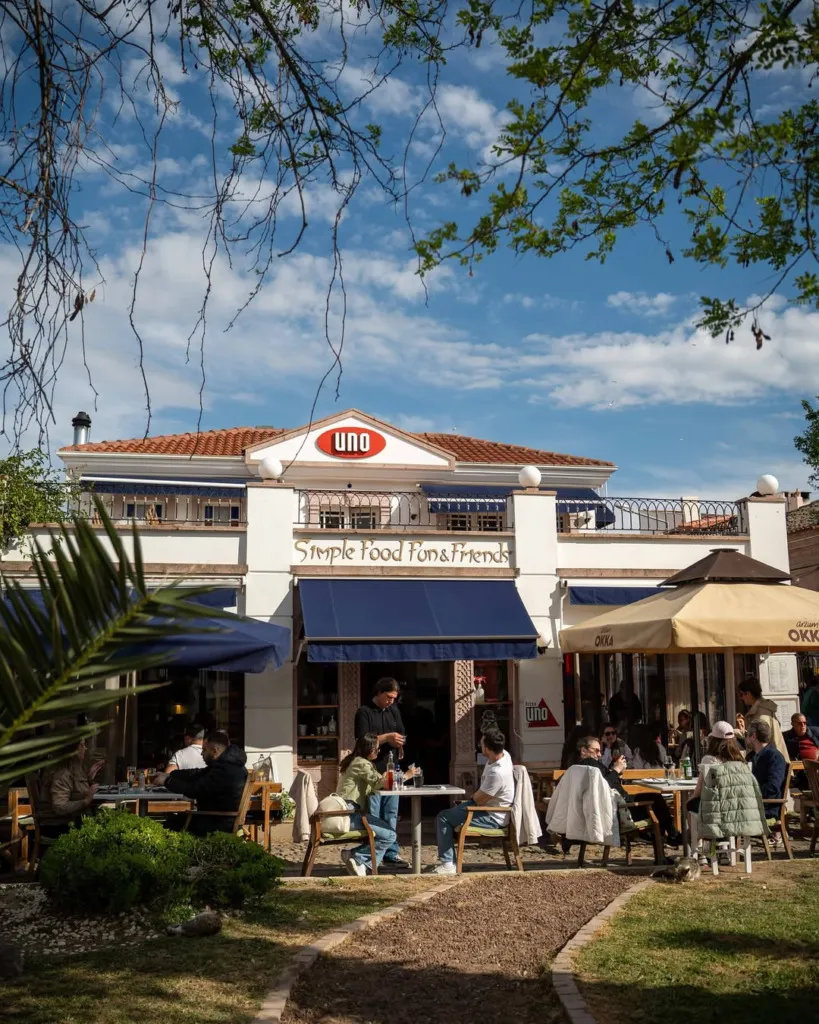 Cunda'da gezilecek yerlerden biri olan, kırmızı kiremit çatılı, beyaz cepheli Uno Simple Food Fun & Friends restoranının canlı dış mekanında, mavi tentelerin altında ve şemsiyelerin gölgesinde yemek yiyen ve sohbet eden insanlarla dolu huzurlu bir bahçe manzarası.