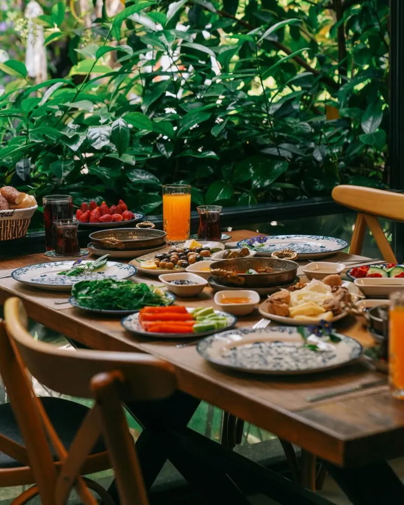 Cunda'da gezilecek yerlerden biri olan yemyeşil bir mekanda, bol çeşitli zeytinler, peynirler, taze yeşillikler, menemen, çilekler, simitler, çay ve portakal suyuyla donatılmış, keyifli bir Türk kahvaltısı sofrası görünüyor.