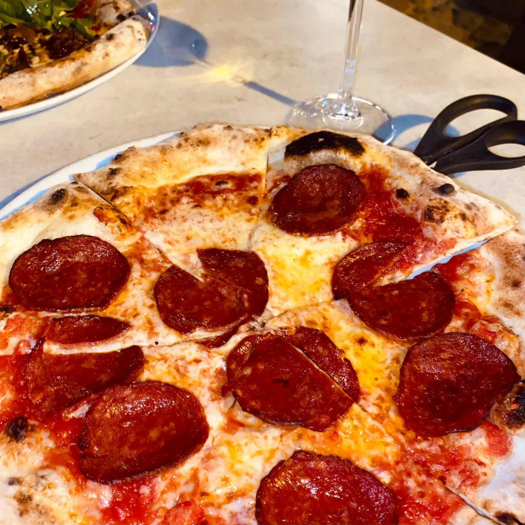 Cunda'da gezilecek yerlerden birinde, üzerinde bolca erimiş mozzarella peyniri ve baharatlı sucuk dilimleri bulunan, ince hamurlu, taze pişmiş, iştah açıcı bir pizza beyaz tabakta servis edilmiş halde, arka planda bulanık bir şarap kadehi ve pizza makası ile görünüyor, bu da ziyaretçilere sunulan lezzetli duraklardan birine işaret ediyor.