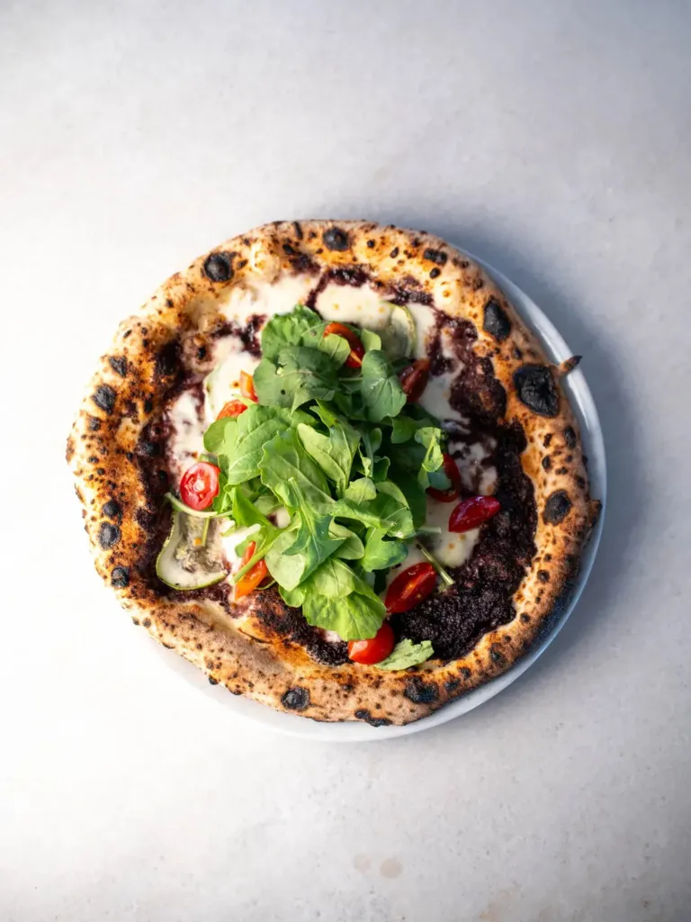 Neapolitan tarzı kabarık ve hafif yanık kenarlı hamuruyla, üzerinde koyu zeytin ezmesi, erimiş beyaz peynir, taze roka yaprakları ve kiraz domates dilimleri bulunan bu iştah açıcı pizza, Cunda'nın zengin mutfağını ve gezilecek lezzet duraklarını çağrıştırıyor.