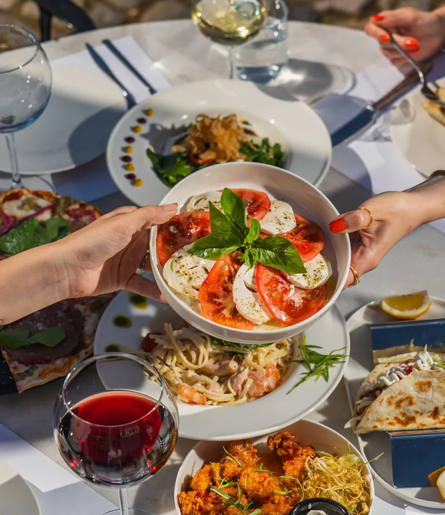 Cunda'nın gezilecek yerlerinden biri olan şirin bir restoranda, güneşli bir masada Caprese salatası, karidesli makarna, pizza ve mezeler gibi çeşitli lezzetler sunulurken, misafirler kırmızı ve beyaz şarap eşliğinde keyifli bir yemeğin tadını çıkarıyor.